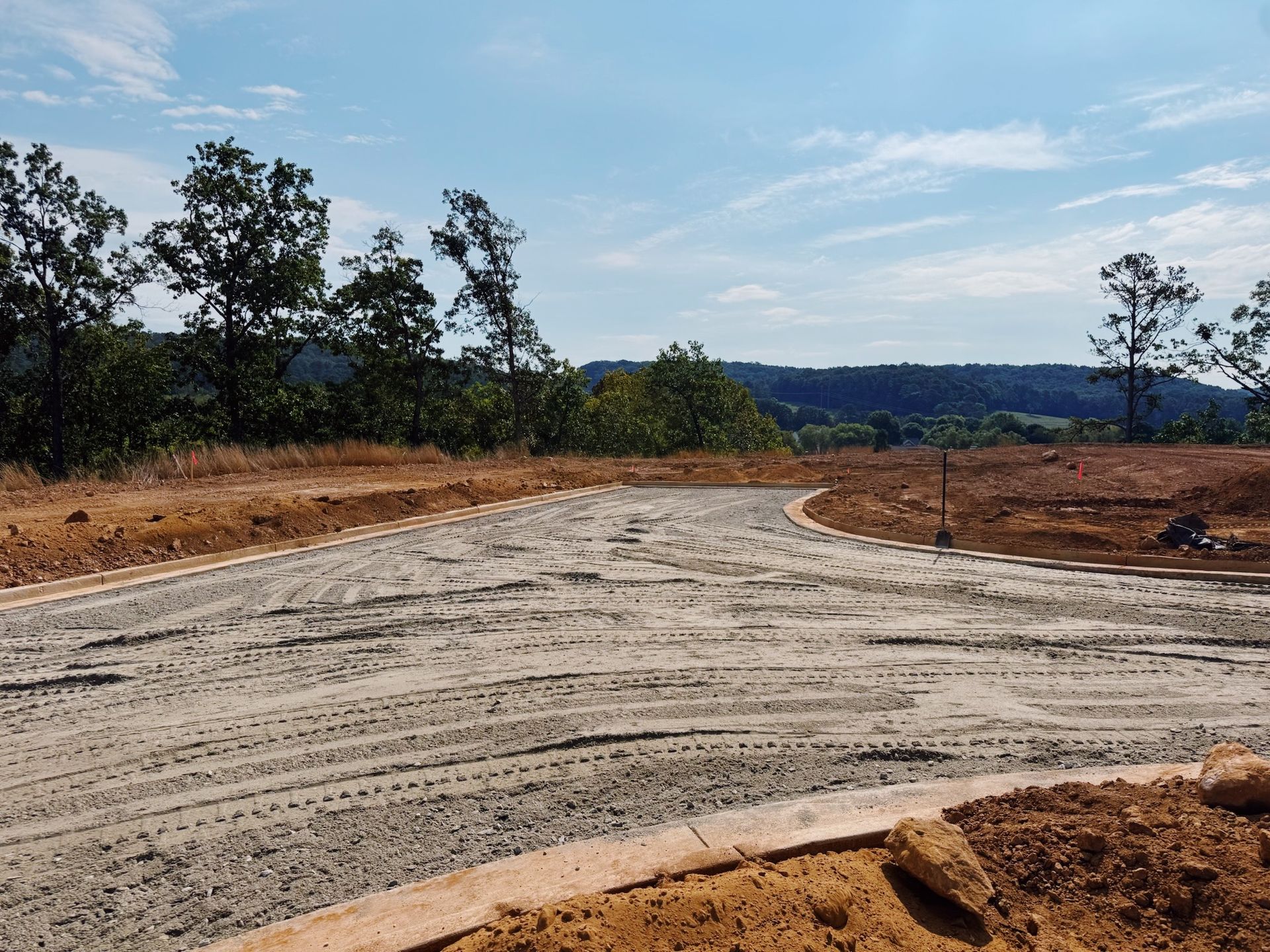 Construction site with gravel road leading toward trees and distant hills under a blue sky.