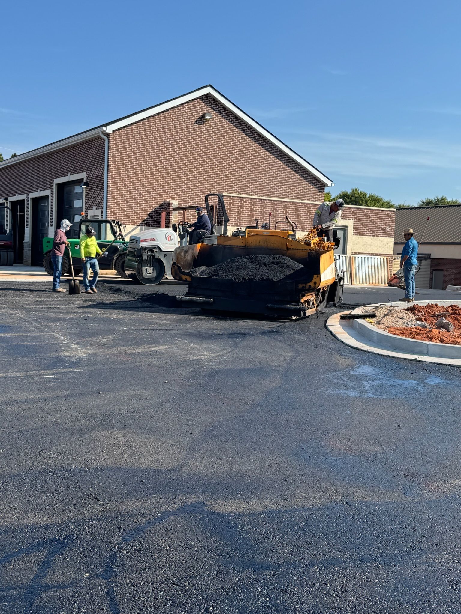 Construction workers paving a parking lot with asphalt. A large paving machine and small tractor are present. Brick building in the background.