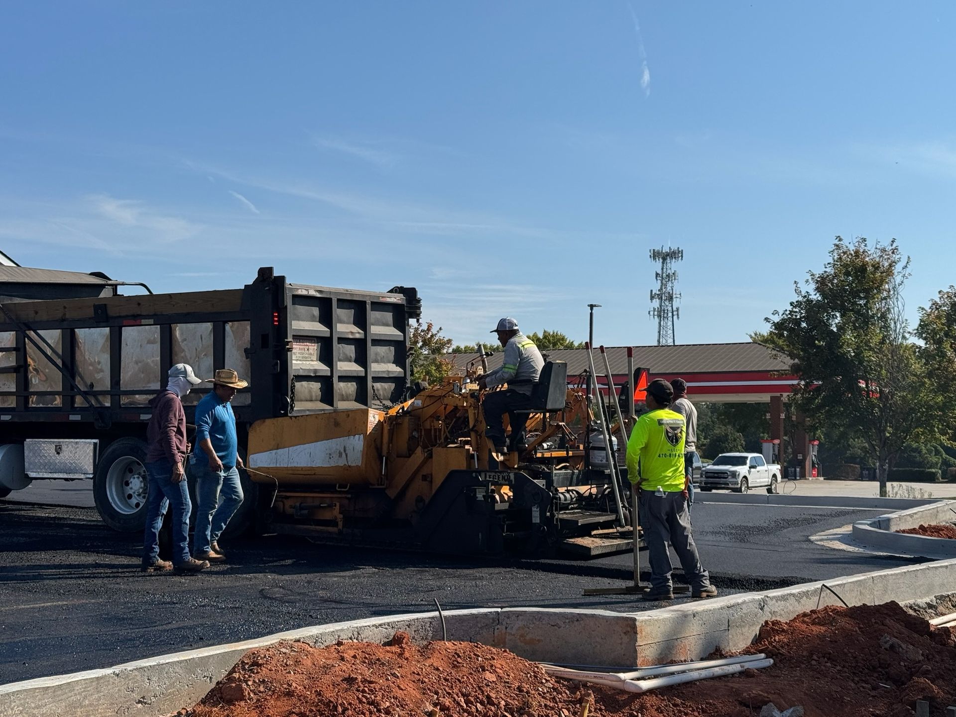 Asphalt paving work: truck, paving machine, workers, freshly laid blacktop, concrete curb, blue sky.