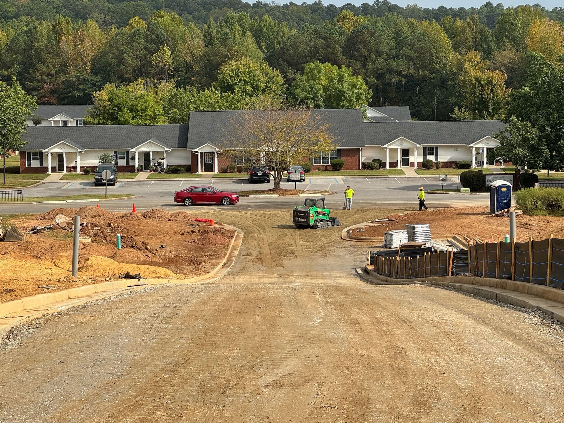 Construction site with grading in foreground and single-story apartments in the background. Workers, a vehicle, and a porta-potty are visible.