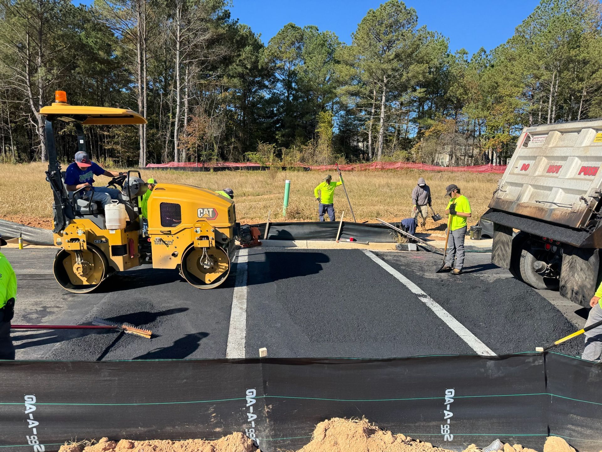 Road construction: Asphalt paving with a roller, workers, and a dump truck on a sunny day.