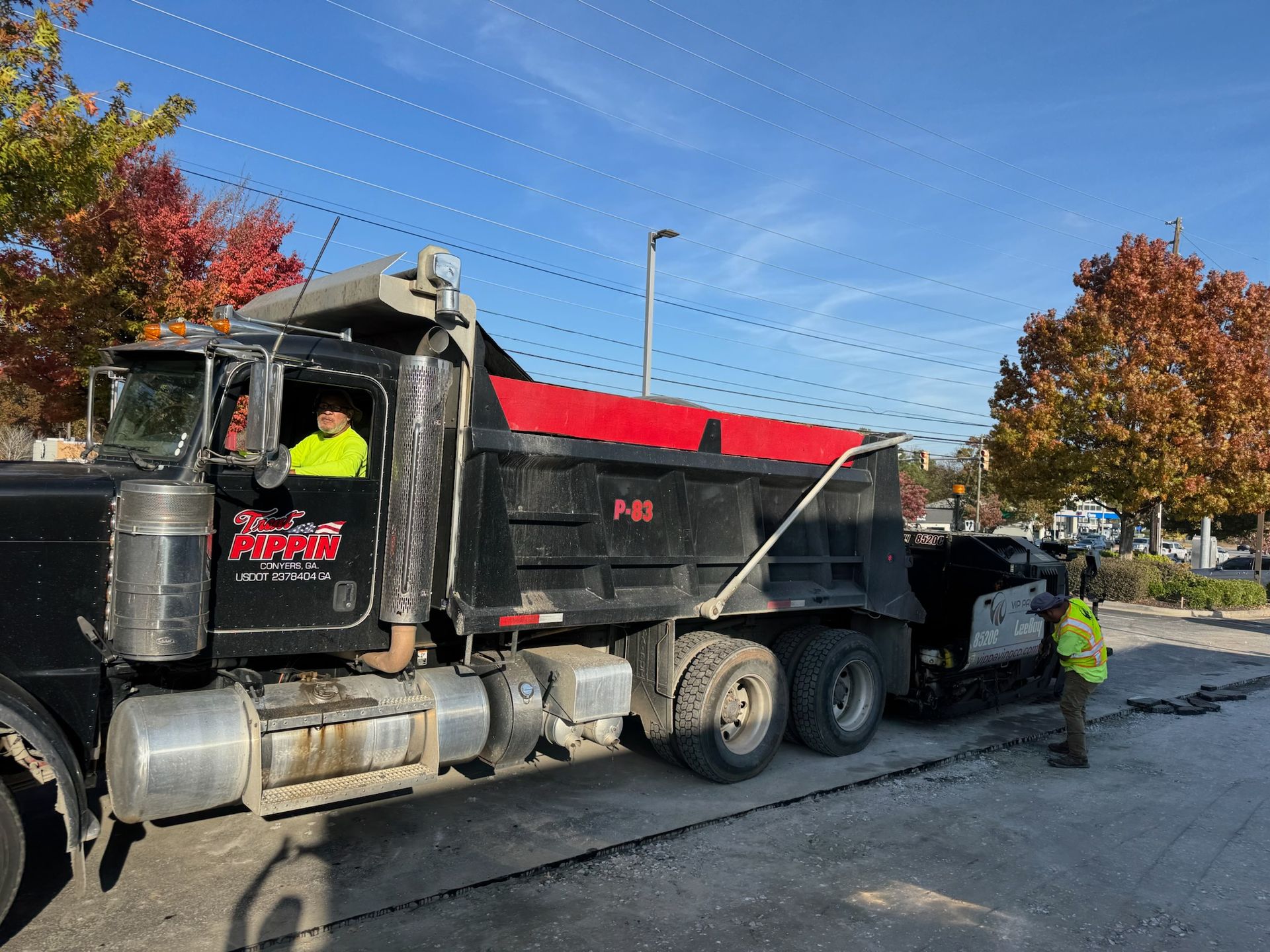 Black dump truck unloading asphalt. Worker in safety vest directs. Sunny day with red and green trees.