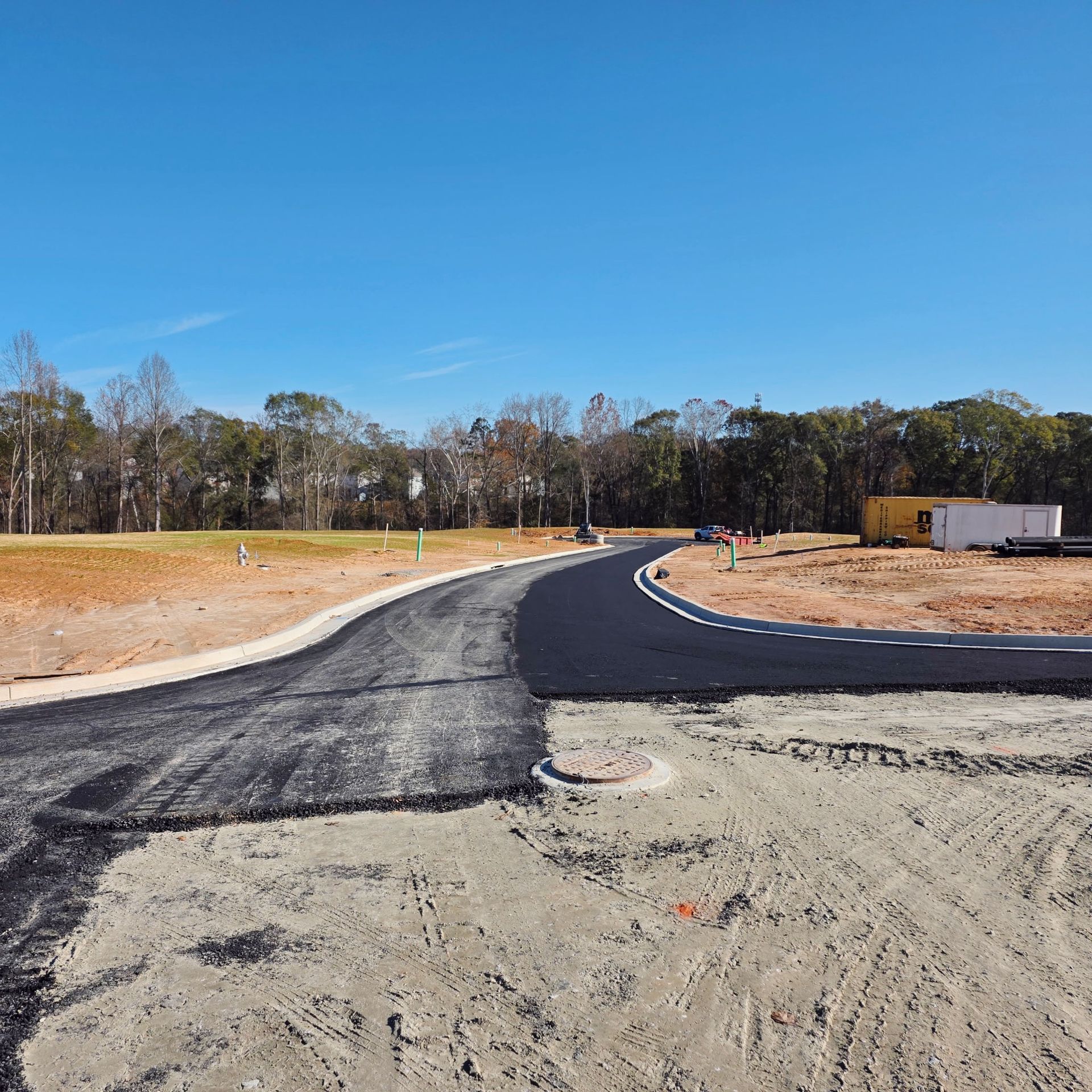 Newly paved road curving through a construction site, with dirt, trees, and blue sky.