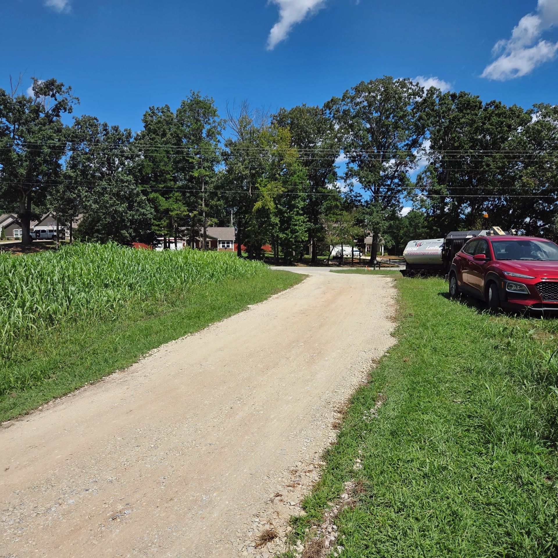 A dirt road leads to a red car and trees on a sunny day with blue sky and cornfield.