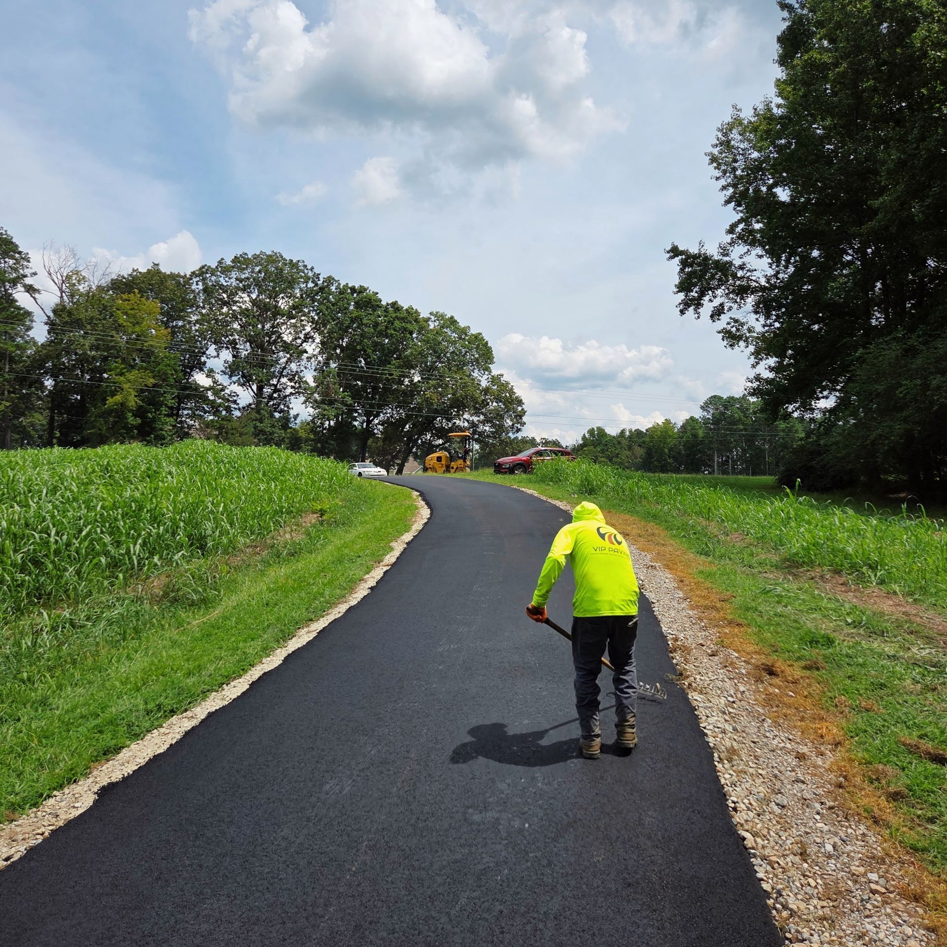 Worker in neon green rakes newly paved asphalt path. Sunny day with trees and tall grass.