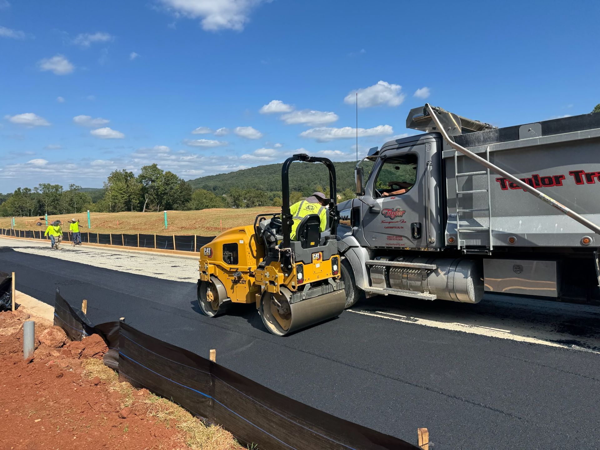 Asphalt paving on a road construction site; roller compacting newly laid asphalt next to a dump truck.
