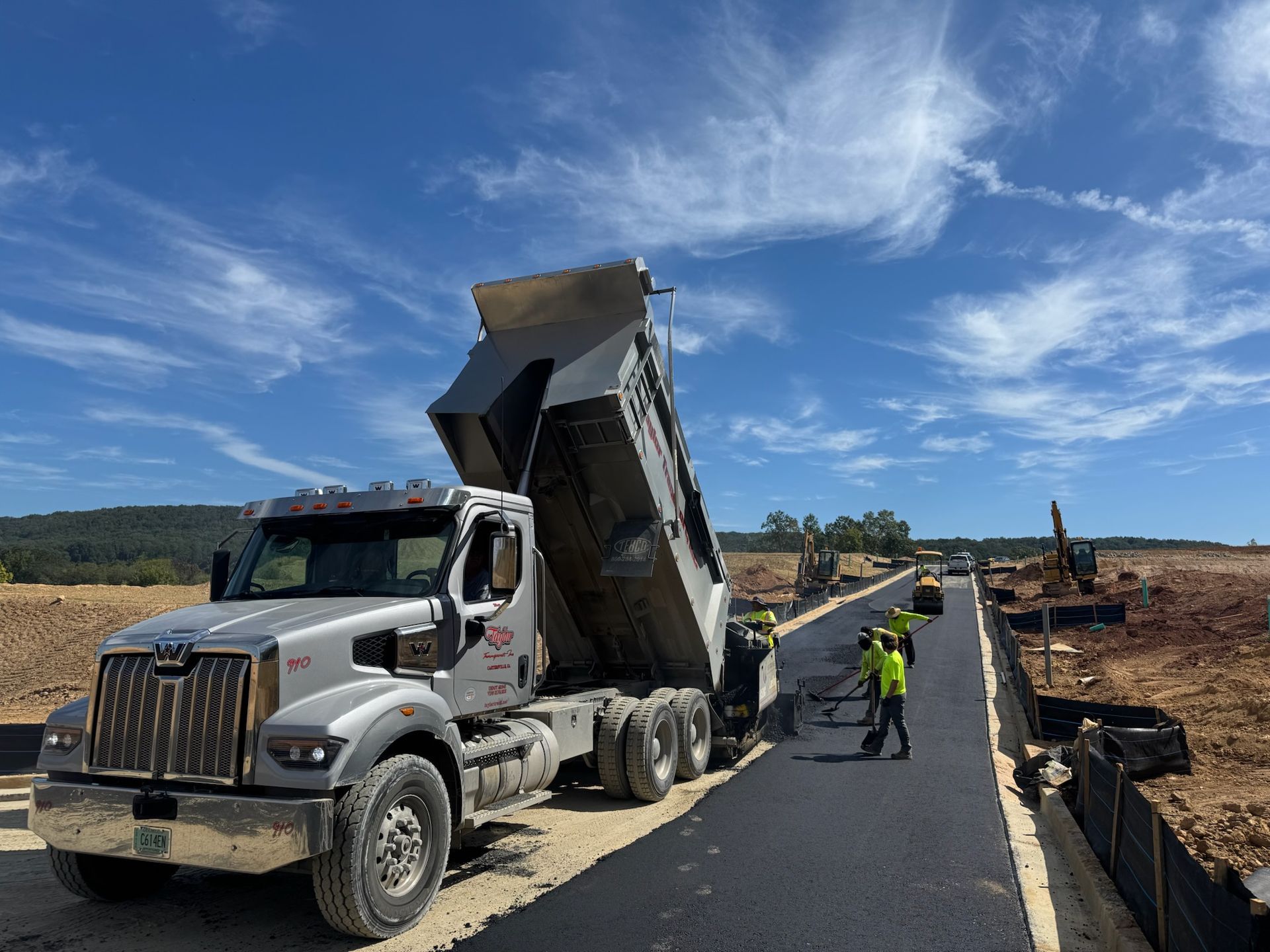 Dump truck unloading asphalt on a newly paved road under a blue sky, construction workers nearby.