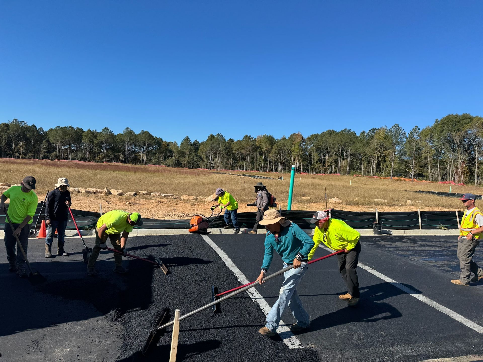 Construction crew laying asphalt on a road under a blue sky. They use tools like rakes and tampers.