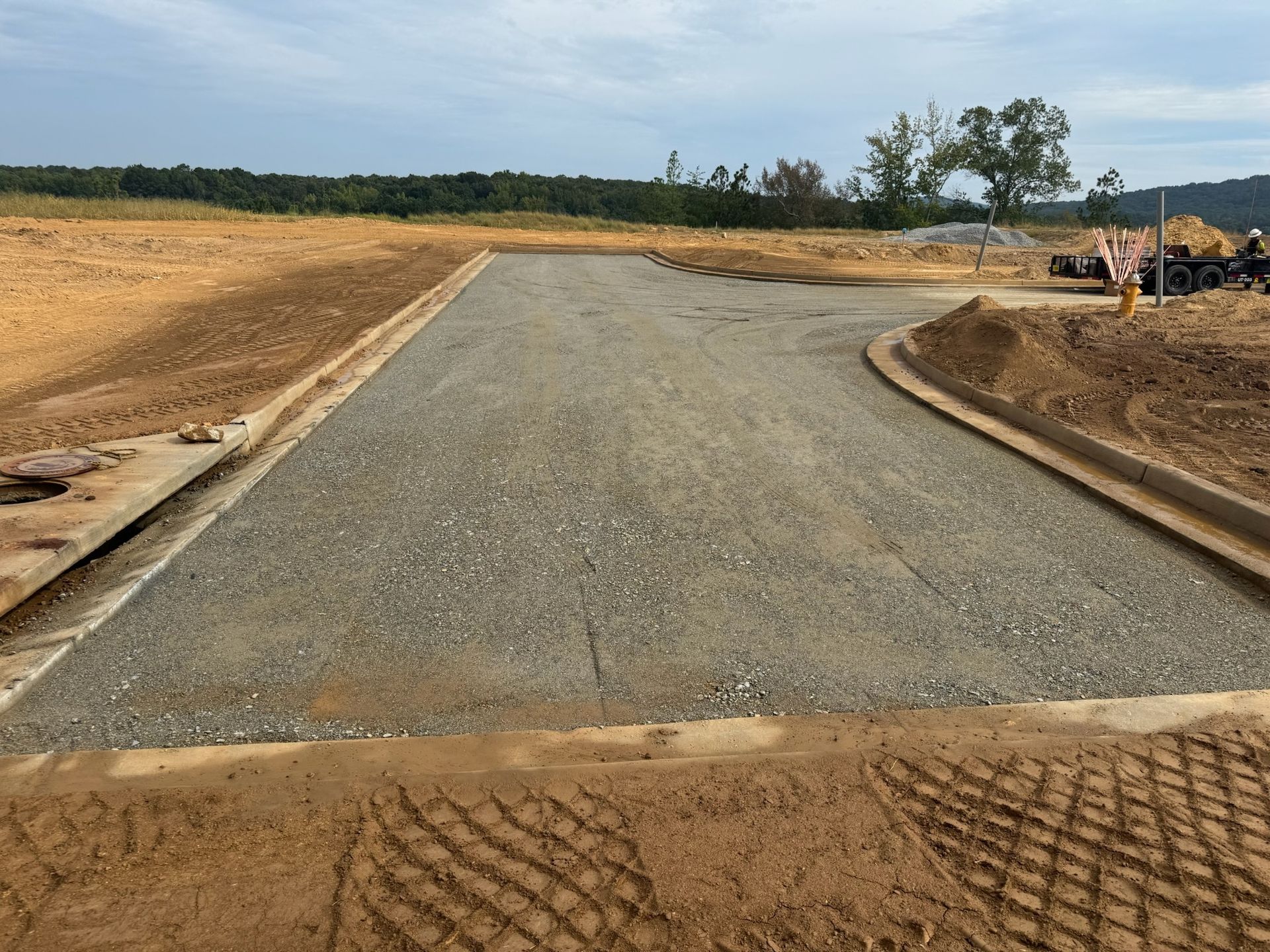 Gravel driveway under construction, with concrete curbs, surrounded by dirt and sparse vegetation.