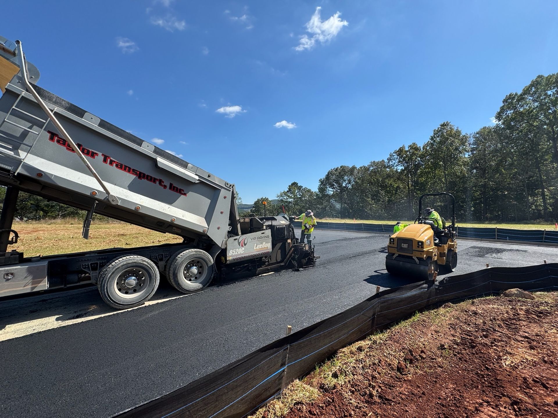 A dump truck unloads asphalt for paving a road. Workers and a roller compact the new surface; sunny day.
