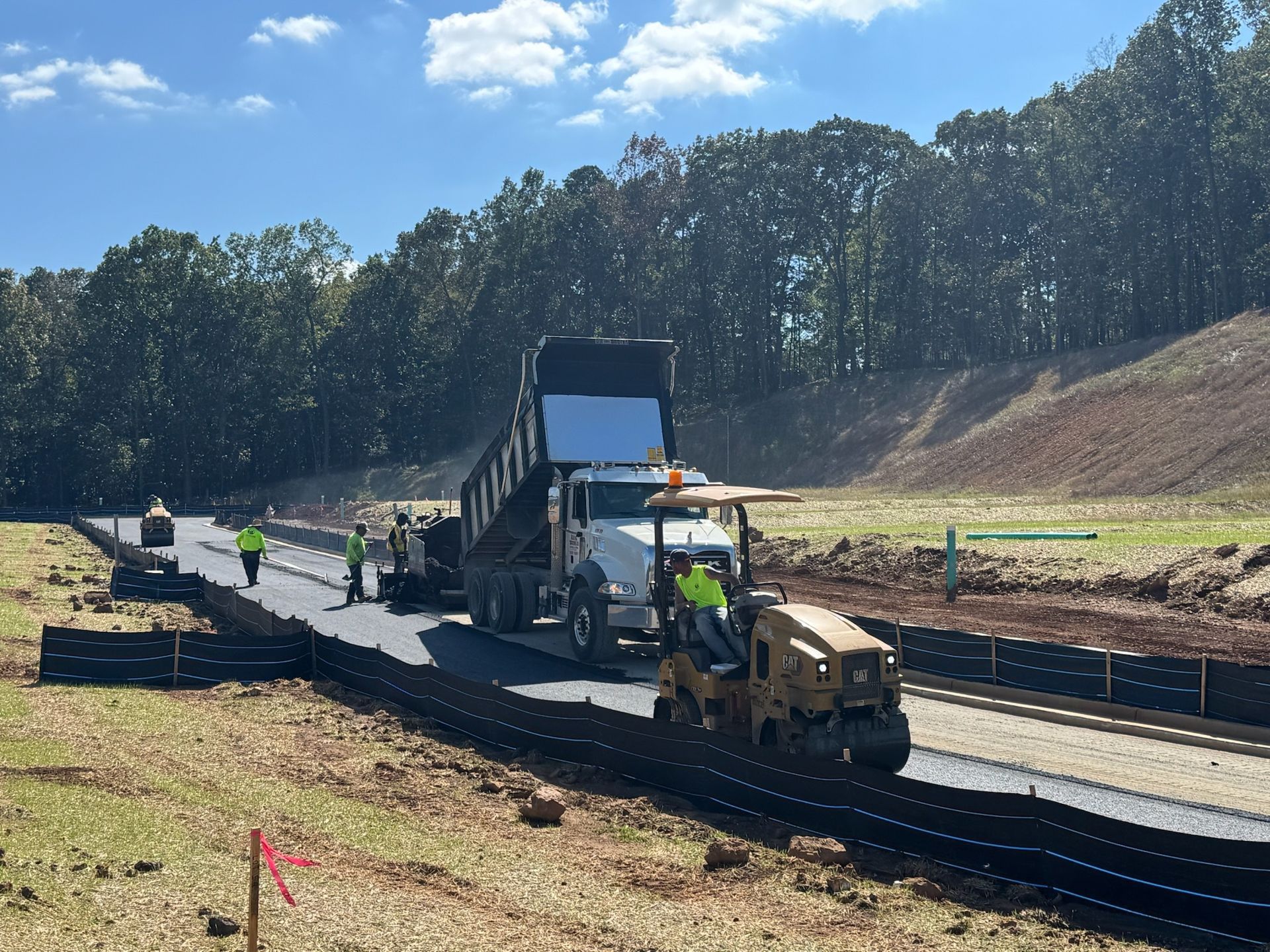 Road construction: dump truck unloading asphalt, workers, roller, blue sky, forest backdrop.
