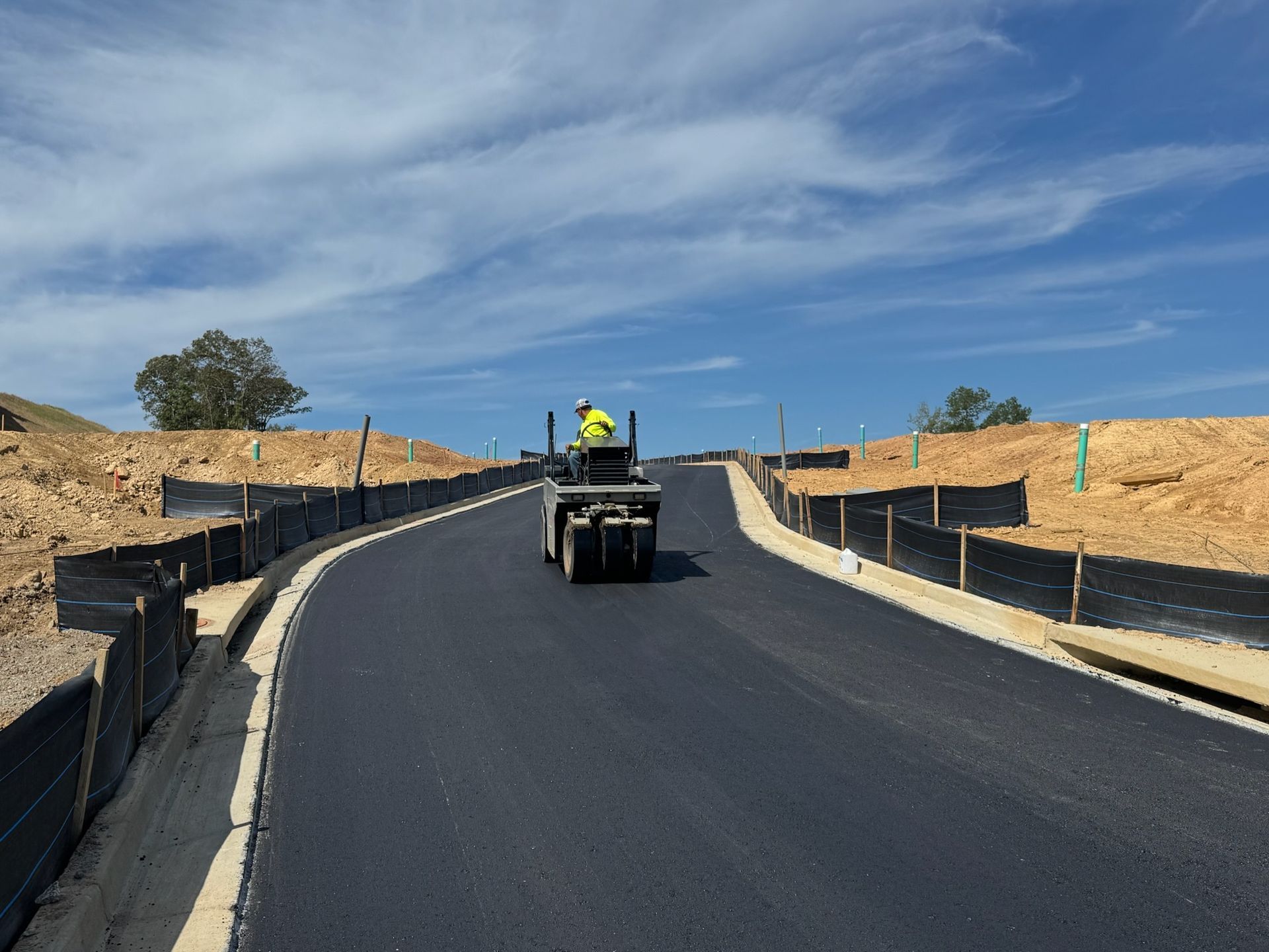 A road paving machine and worker laying asphalt on a curved road under a blue sky.
