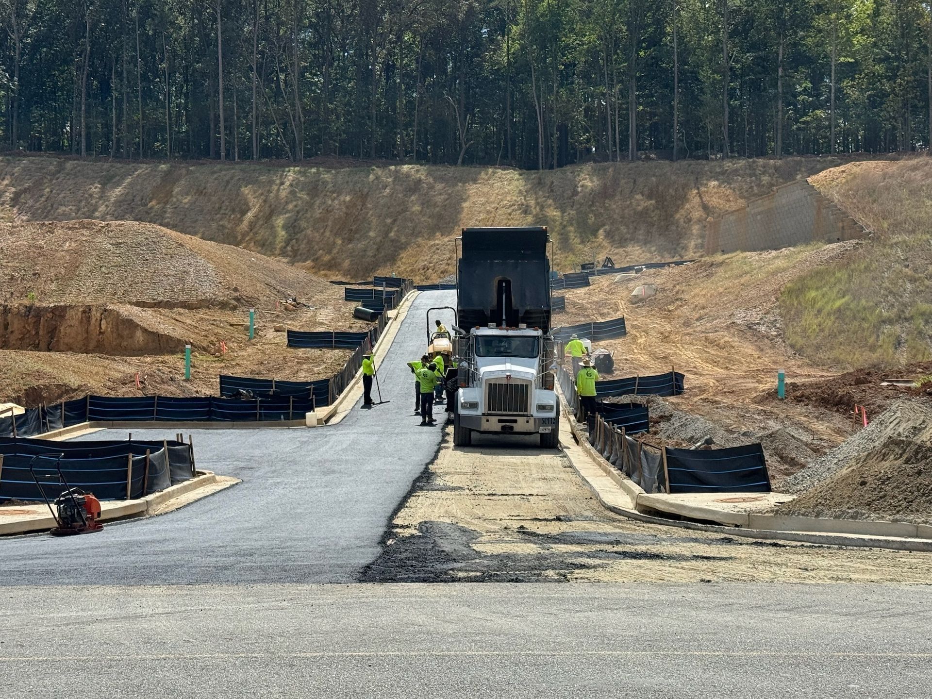 Construction site with a dump truck unloading gravel onto a paved road. Workers in yellow vests.