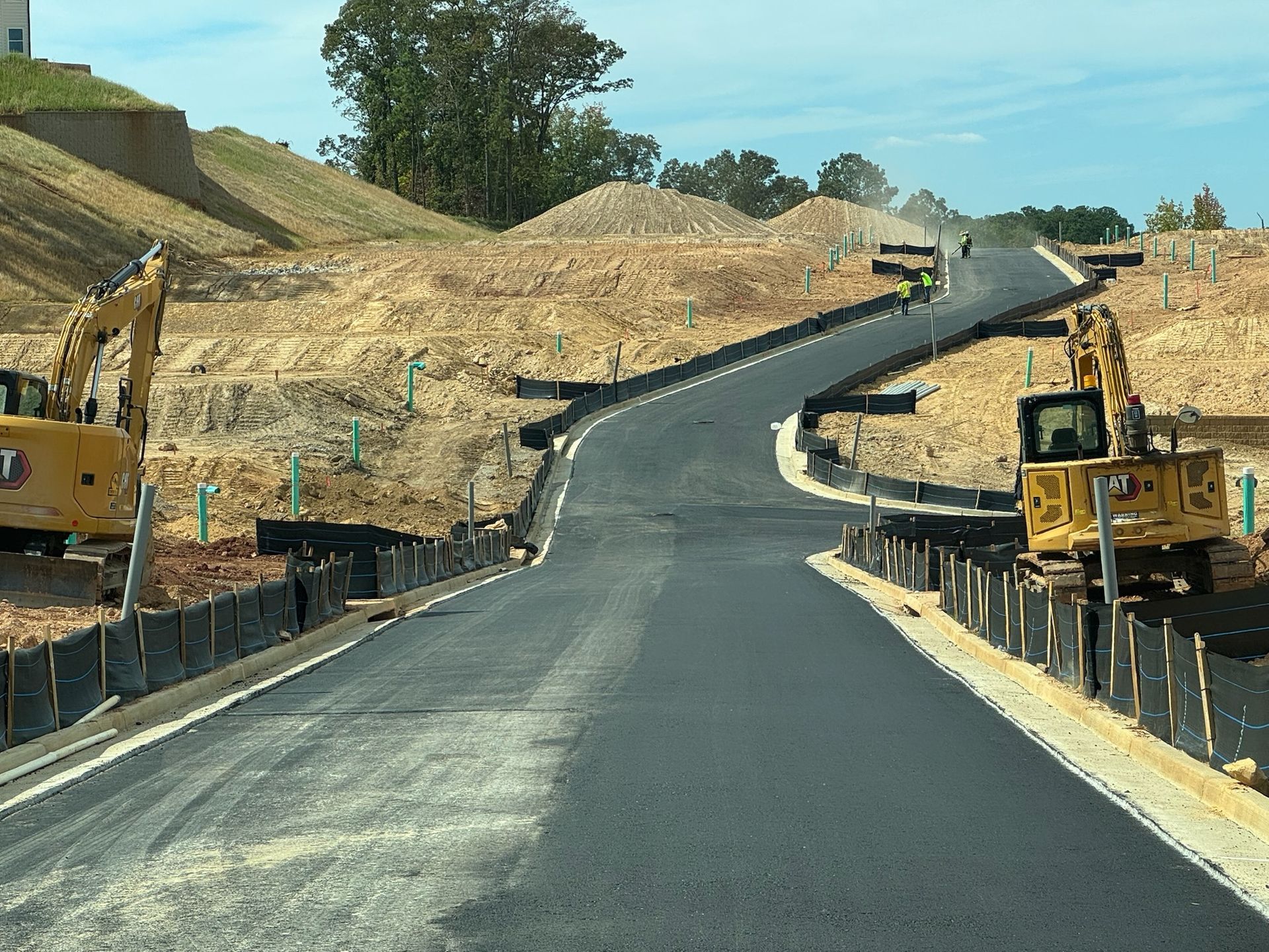 Construction site with newly paved road winding uphill, flanked by excavators and erosion control barriers.