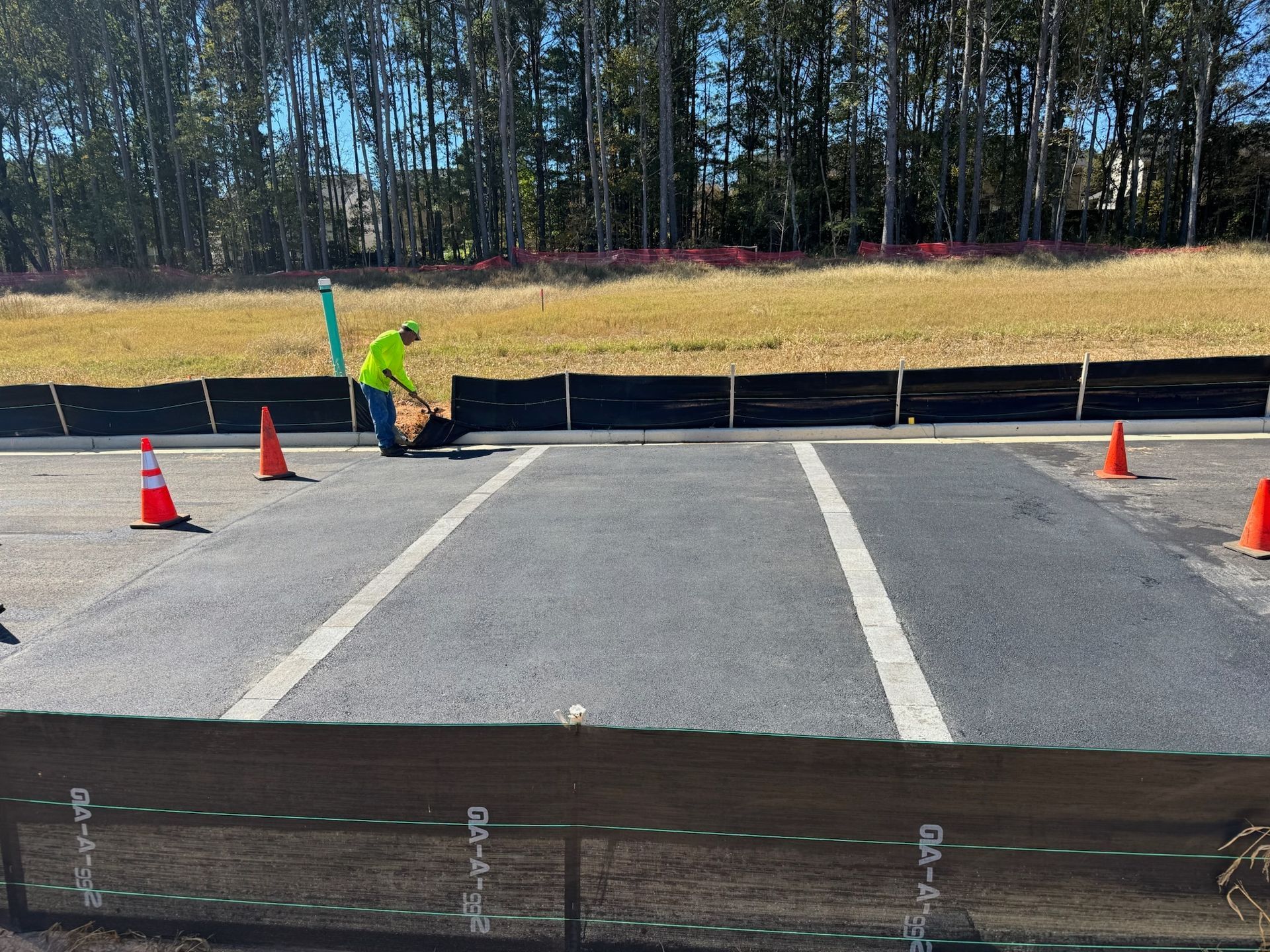 Person in neon green vest working on asphalt road surface, bordered by cones and silt fence.