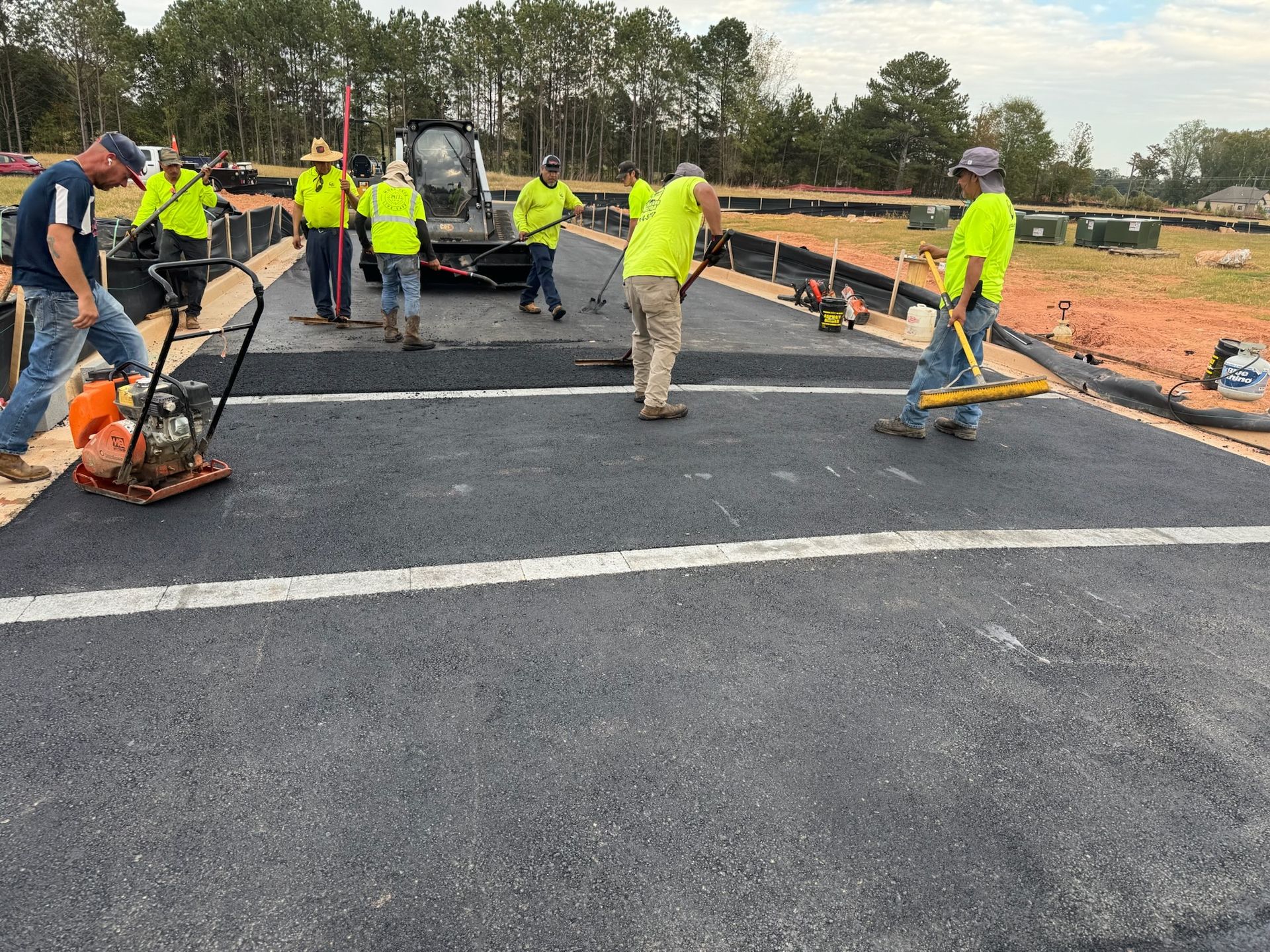 Construction workers paving asphalt road, using equipment and tools, under a partly cloudy sky.