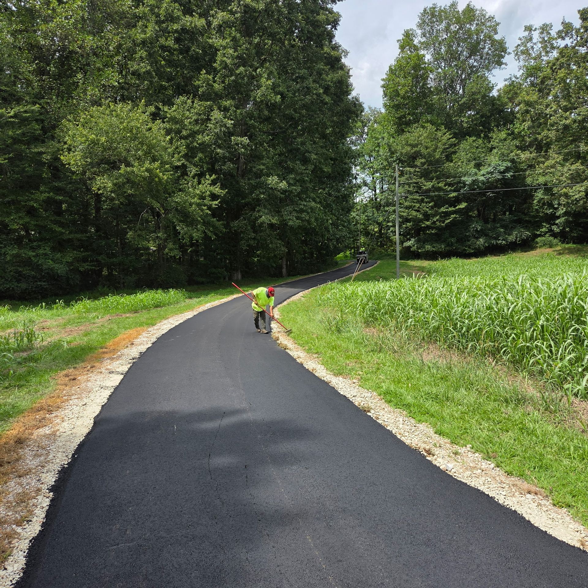 A paved path curves through a grassy area, with trees lining either side. A person in yellow works on the path.