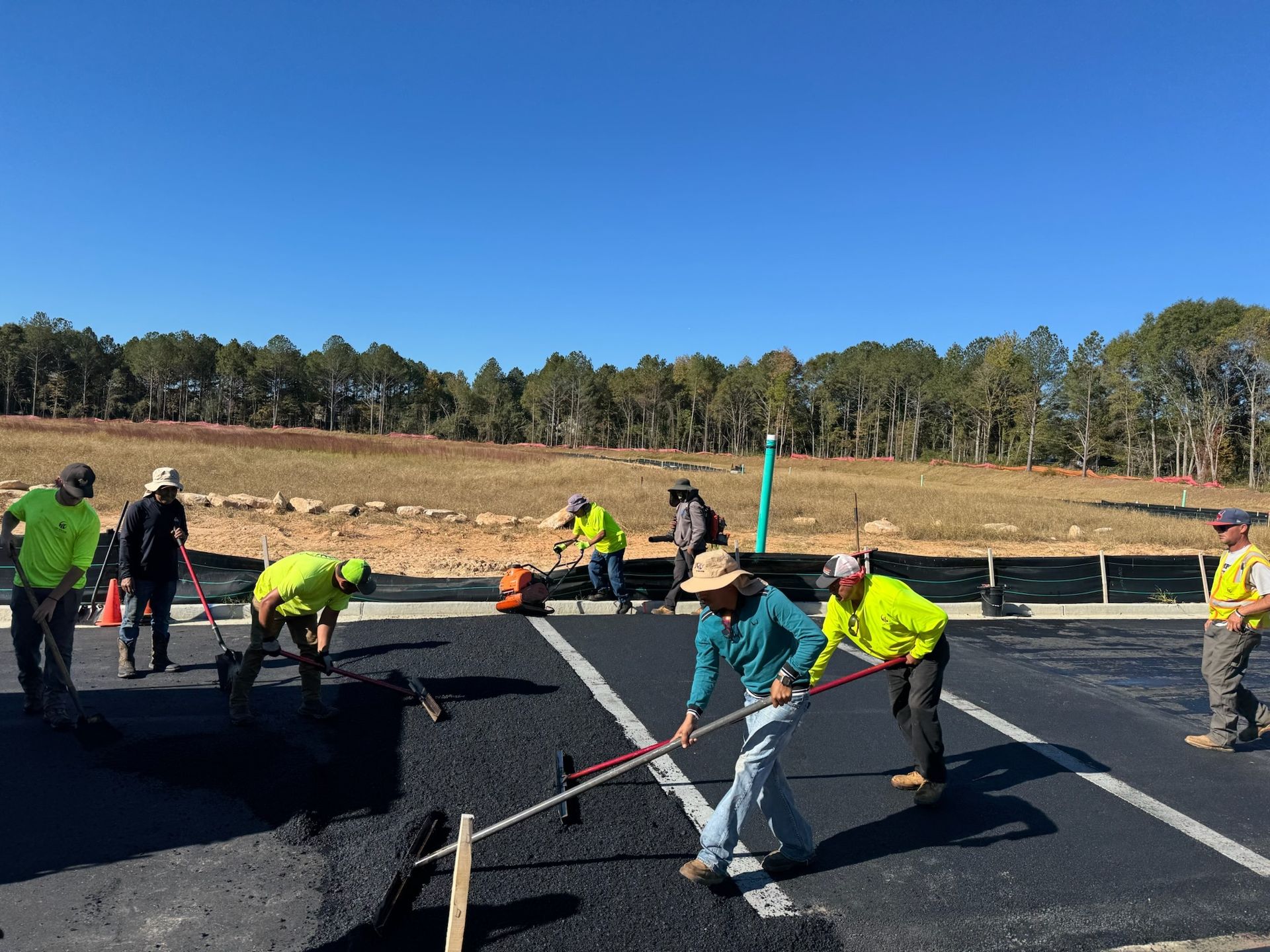 Construction workers paving a parking lot. Asphalt being laid with various tools under a clear blue sky.