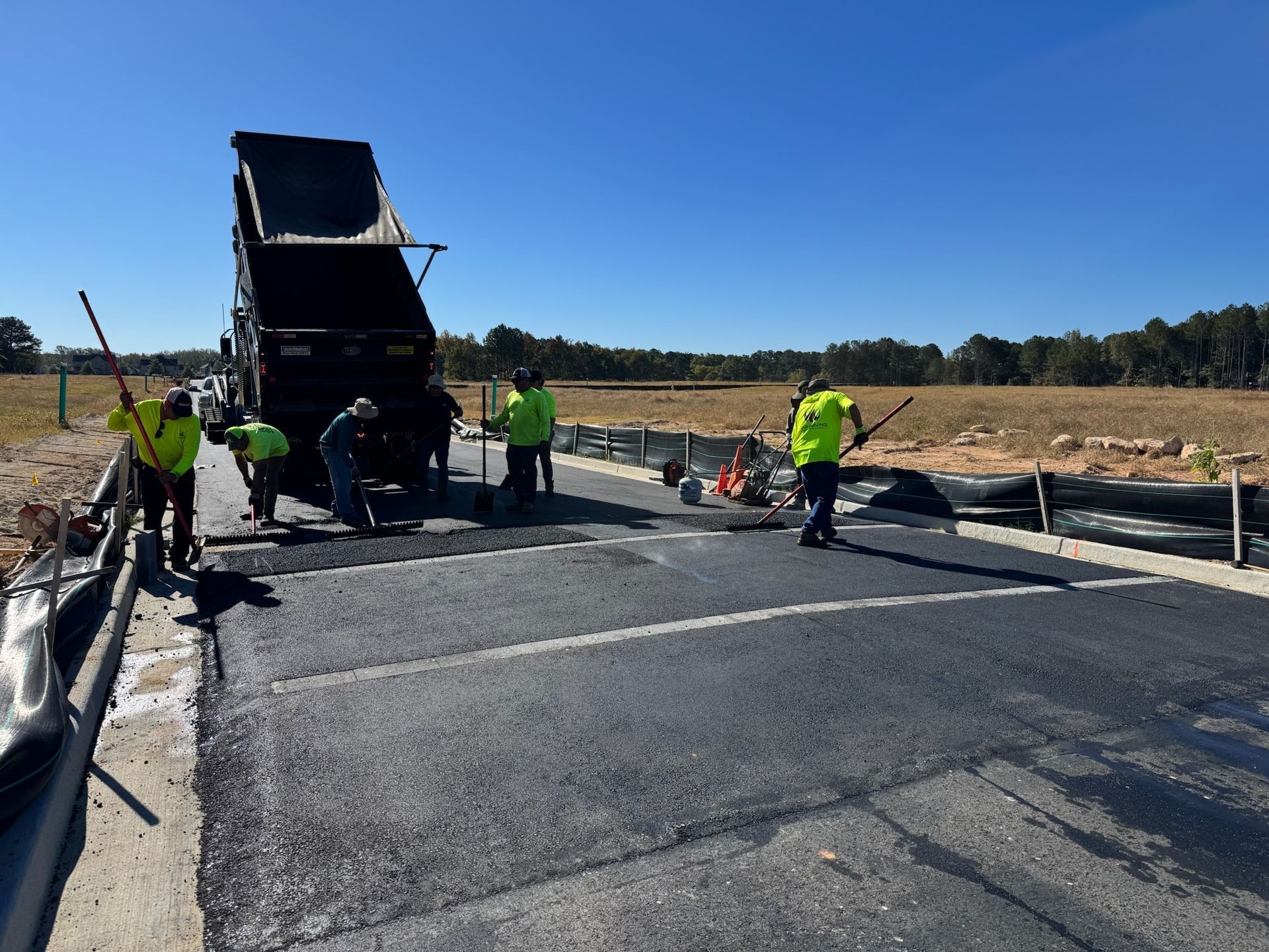 Road construction: Workers in safety vests paving asphalt on a sunny day, dump truck unloading.