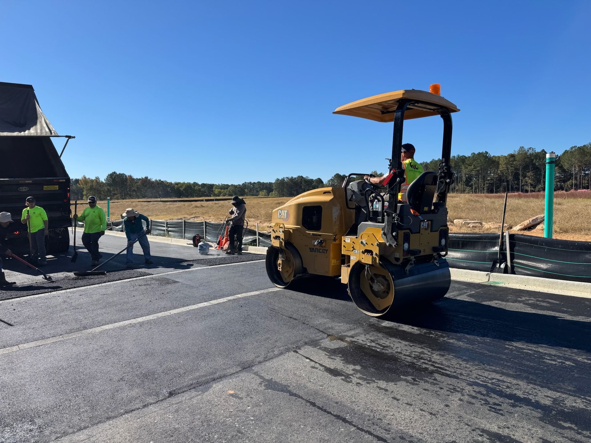Road construction site with a yellow steamroller, workers paving road on a sunny day.