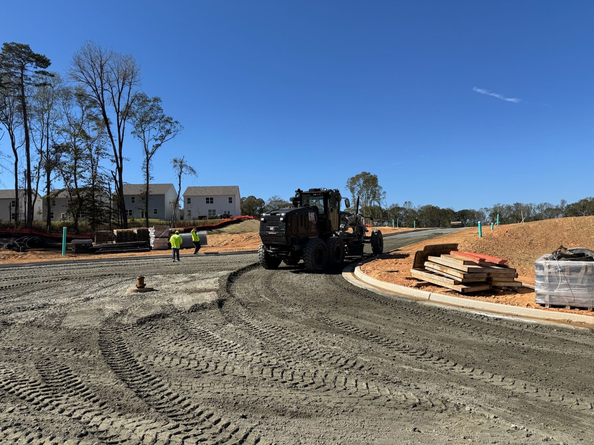 Construction site with a grader, workers, and piles of materials under a blue sky.