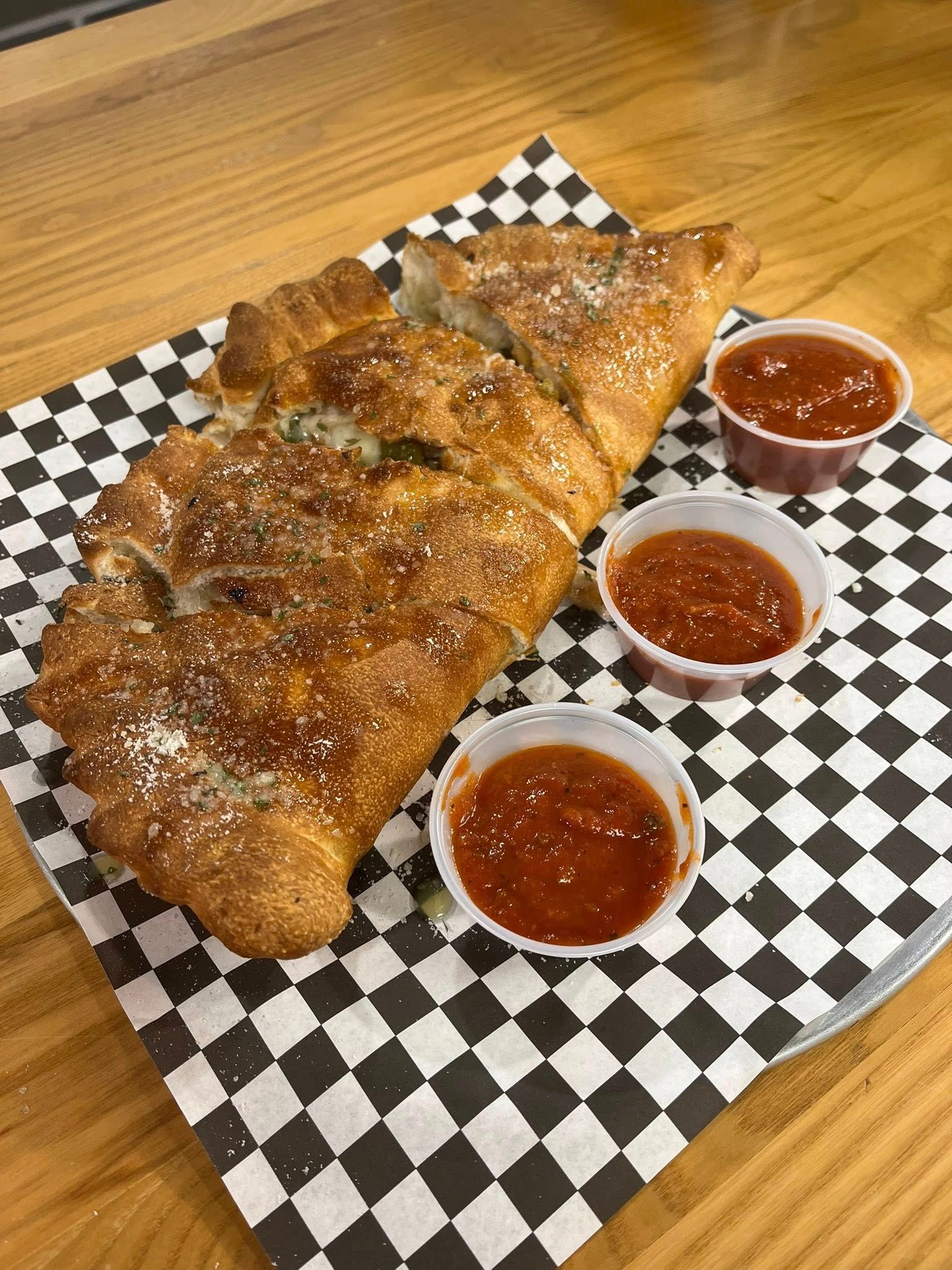Calzone with tomato sauce in small cups on a checkered paper, served on a wooden table.