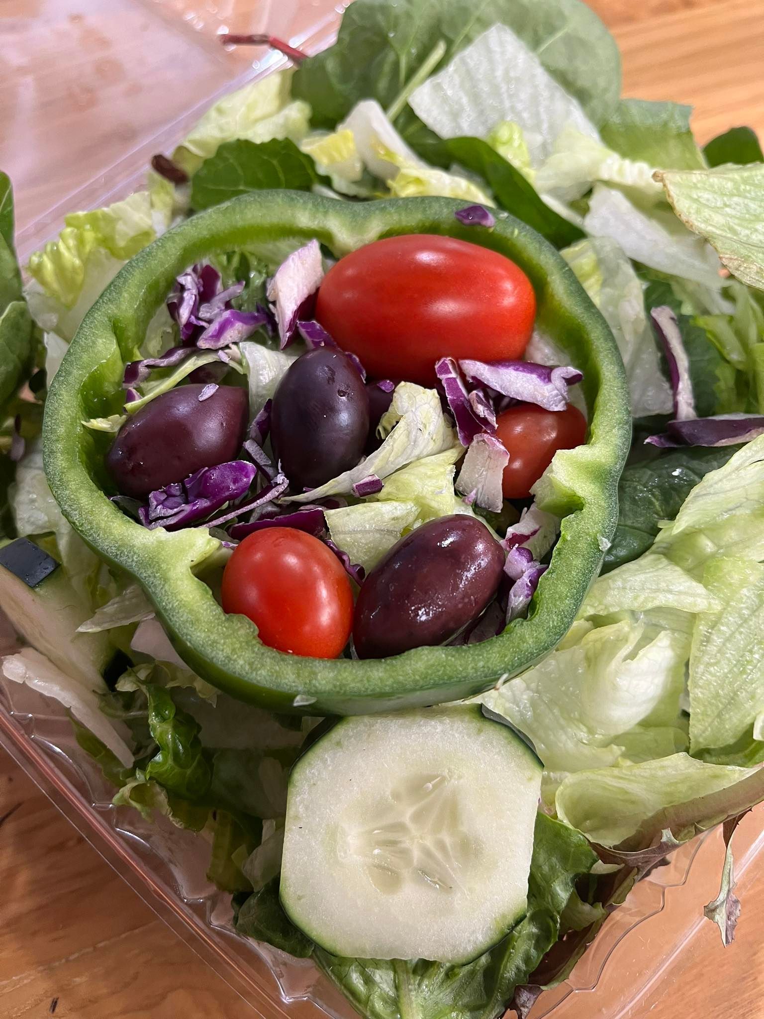 Salad with olives, tomatoes, and purple cabbage inside a green pepper ring, on a bed of lettuce, with a cucumber slice.