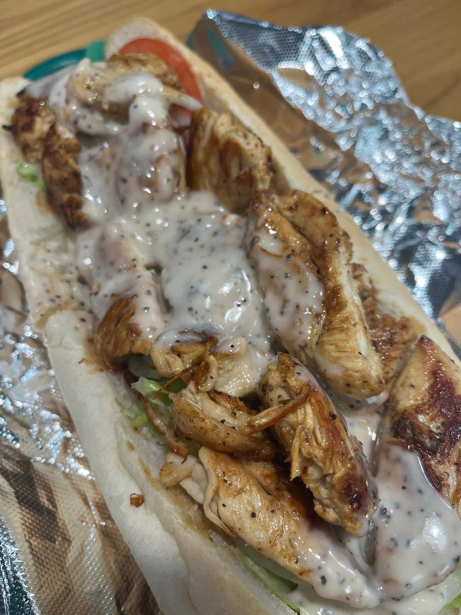 A close-up of a chicken sandwich on a sub roll, topped with white sauce. The sandwich rests on foil.