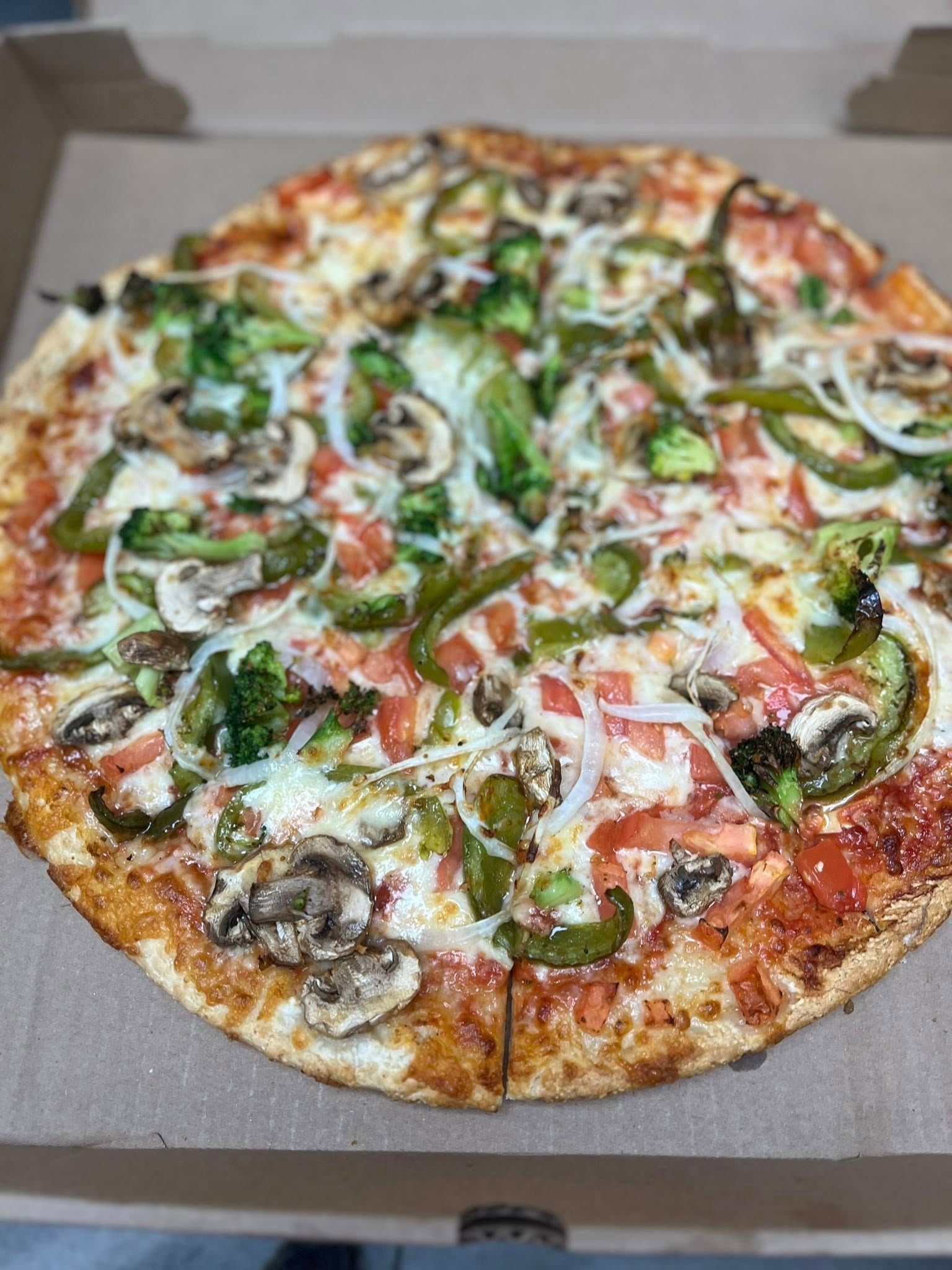Vegetable pizza in a cardboard box, with green peppers, broccoli, onions, and mushrooms on a tomato sauce and cheese base.