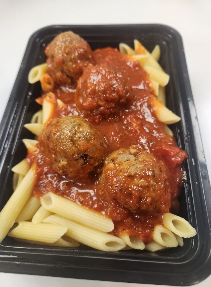 Pasta with meatballs in a black takeout container. Penne pasta is topped with tomato sauce and three meatballs.