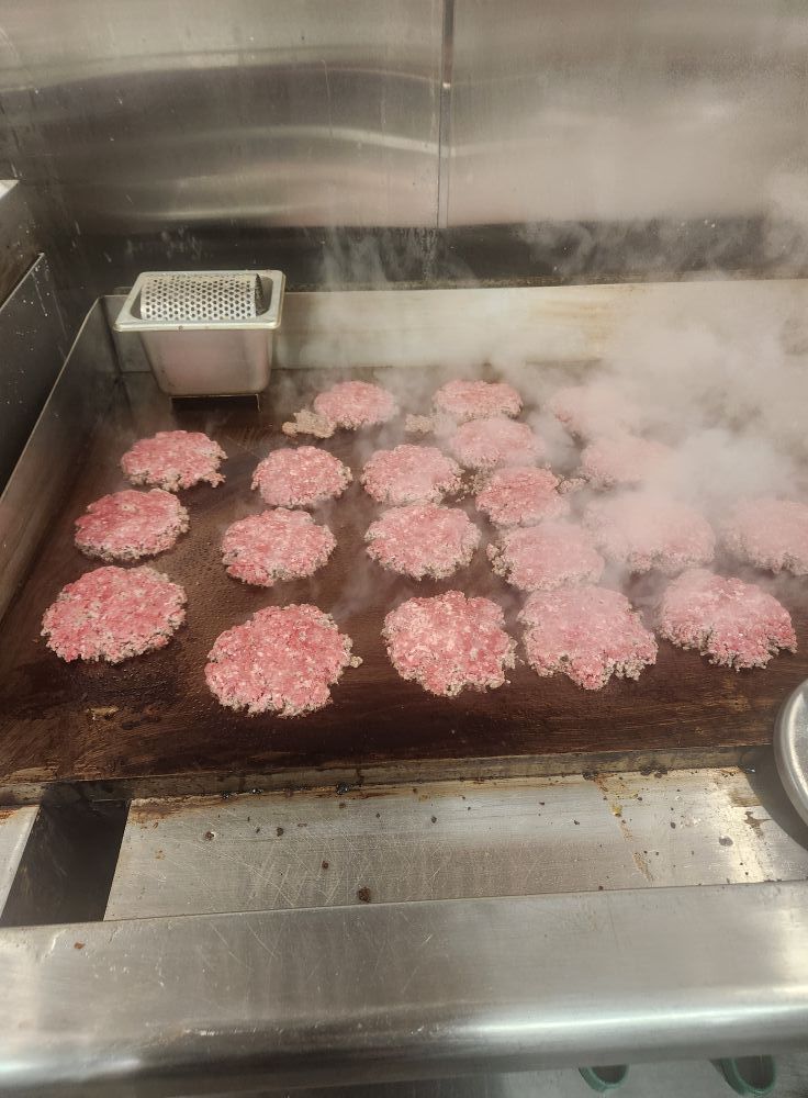 Raw hamburger patties cooking on a hot griddle, creating steam. The setting appears to be a commercial kitchen.