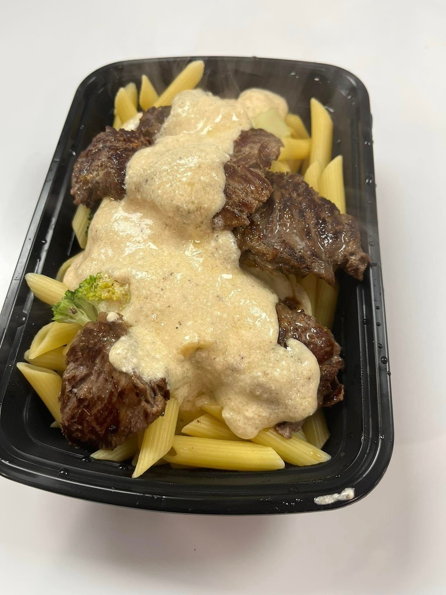 Steak and penne pasta meal in a black container, covered in creamy sauce. Includes broccoli.