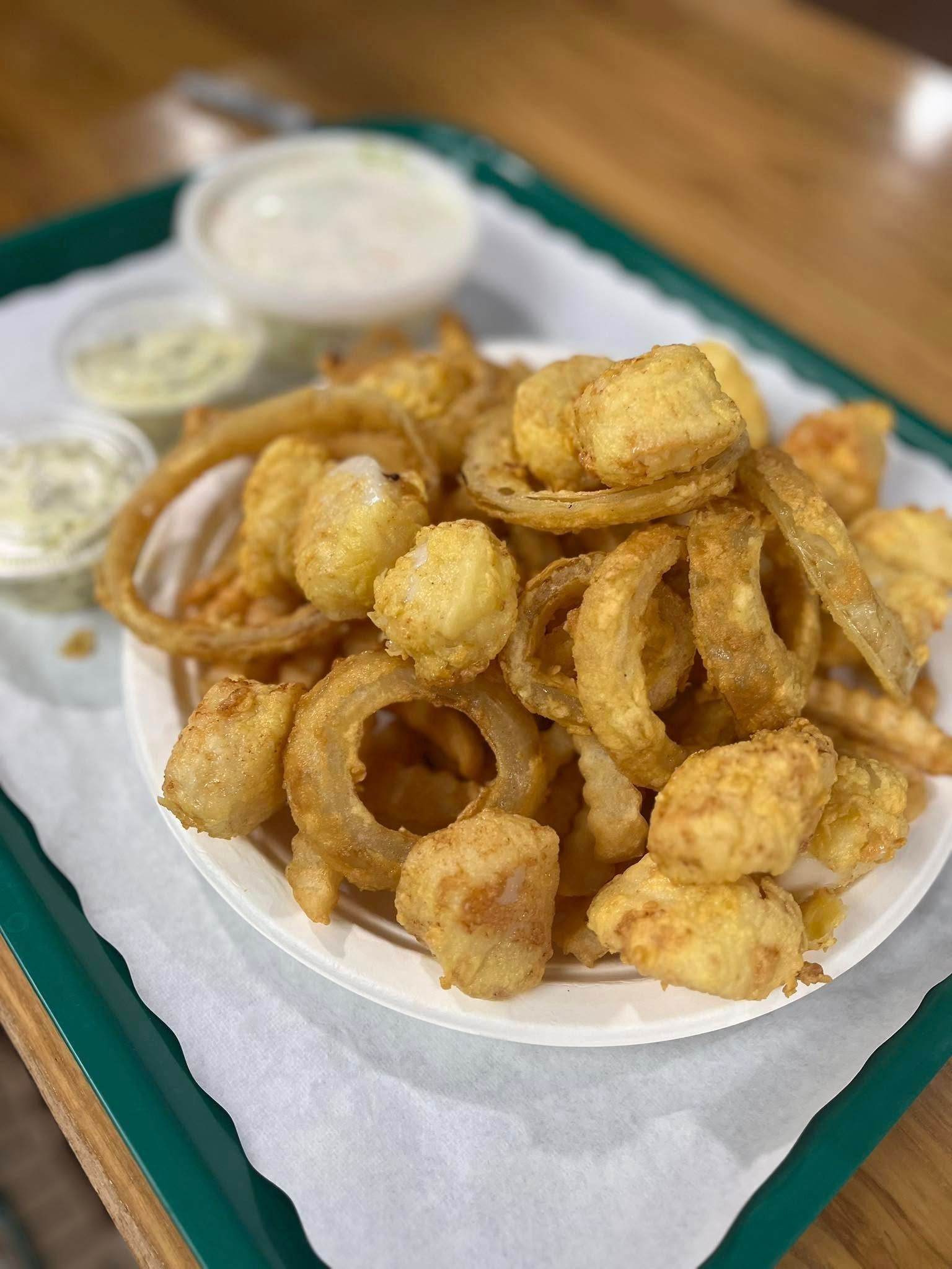 A plate of fried seafood, including onion rings and fried clam pieces, with three small cups of dipping sauce on a green tray.