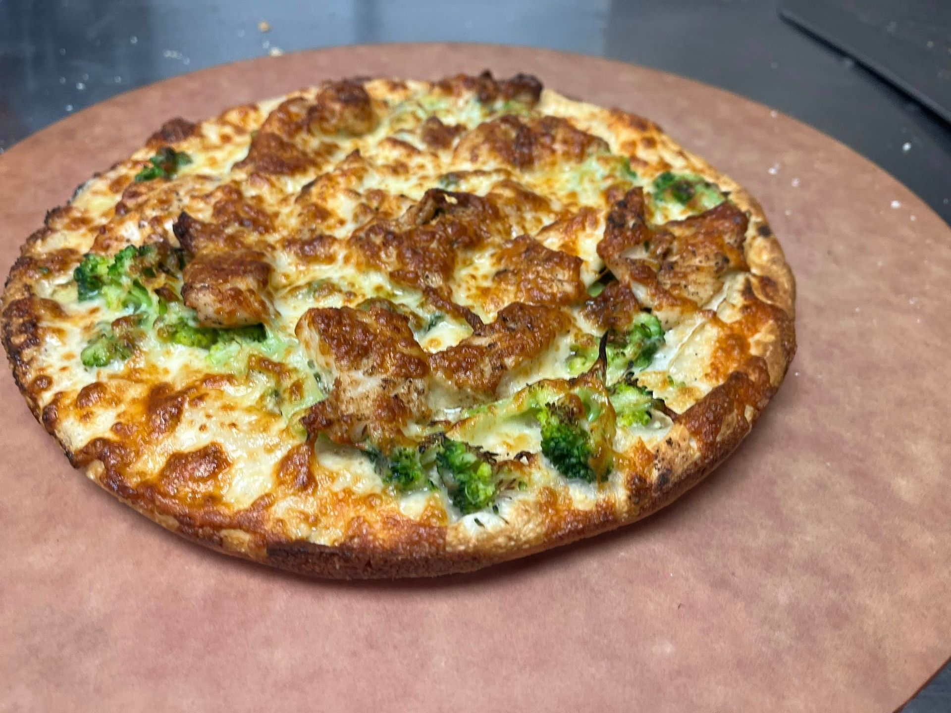Pizza with chicken and broccoli on a round wooden pizza peel. The crust is golden brown and the cheese is melted.