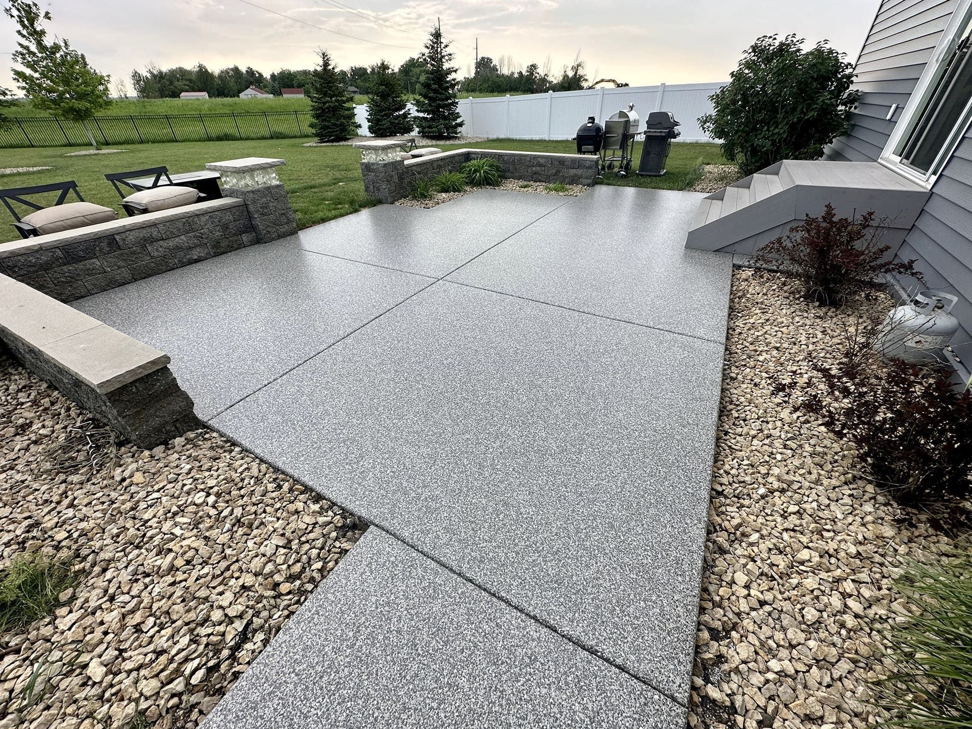 Outdoor stone patio and walkway with gravel borders, benches, and two people near a lake at sunset
