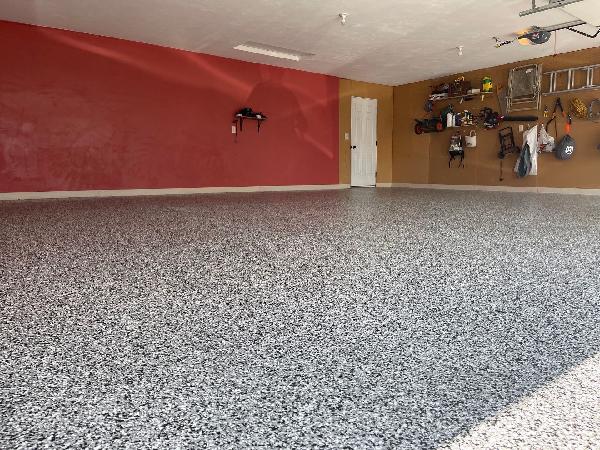 A garage with a speckled gray floor, red and tan walls, and tools hanging on one side.