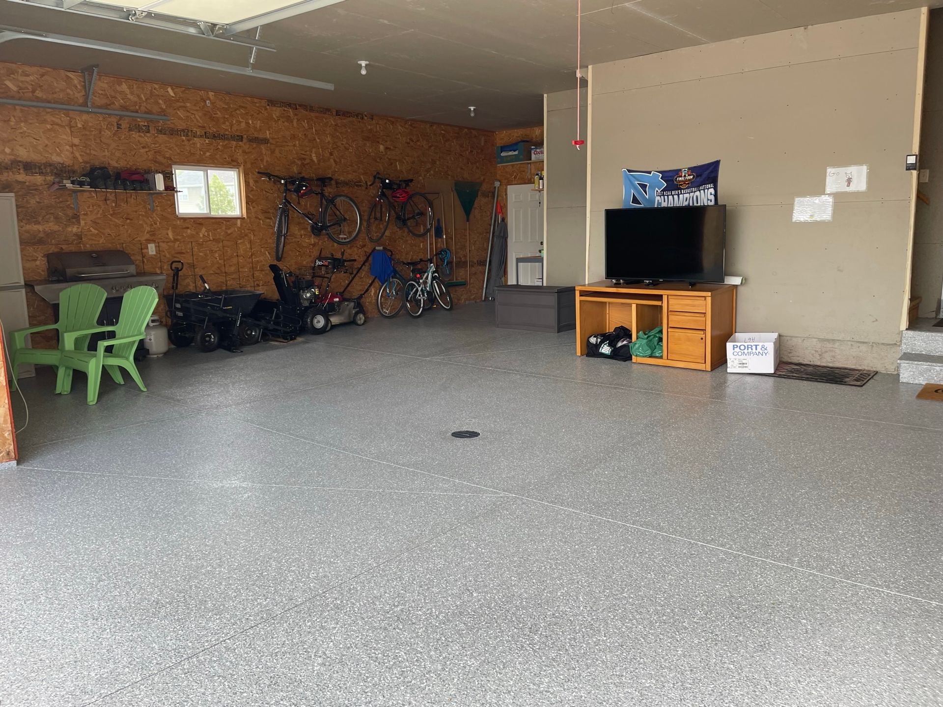 A garage interior with epoxy floor, holding bicycles and tools. Two green chairs sit near a wall with exposed wood. A TV is on a wooden stand.