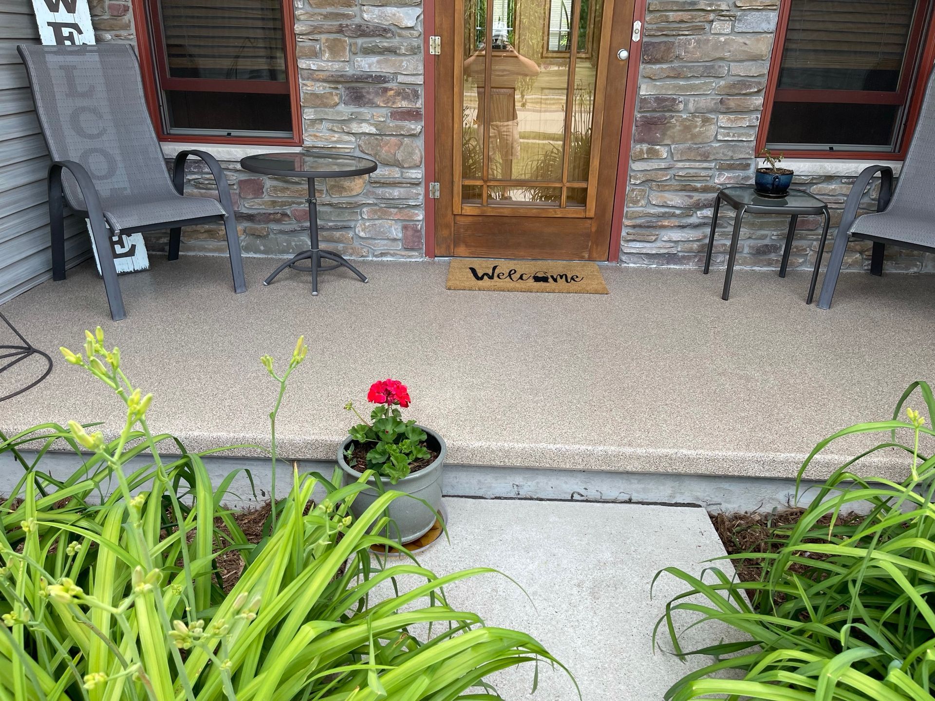 A front porch with stone facade and a clear-paneled door, two chairs, and a welcome mat. A flower pot sits in front.