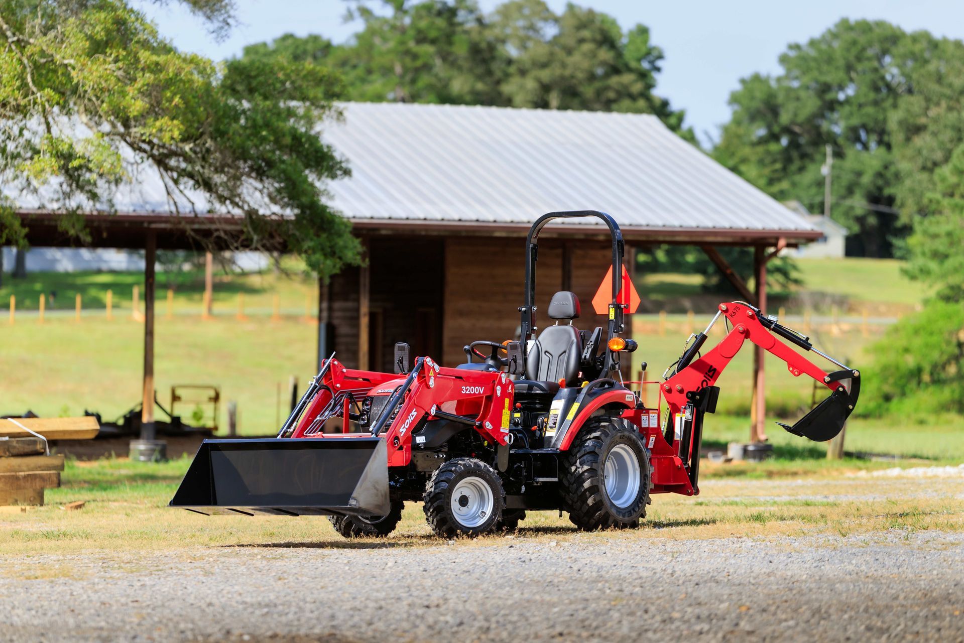 Red tractor with front loader and backhoe on a gravel surface near a wooden building.