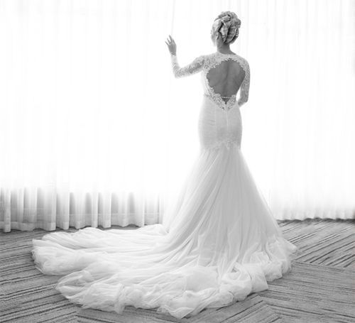 A black and white photo of a bride in a wedding dress standing in front of a window.