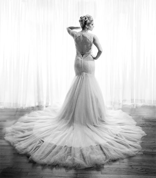 A black and white photo of a bride in a wedding dress standing in front of a window.