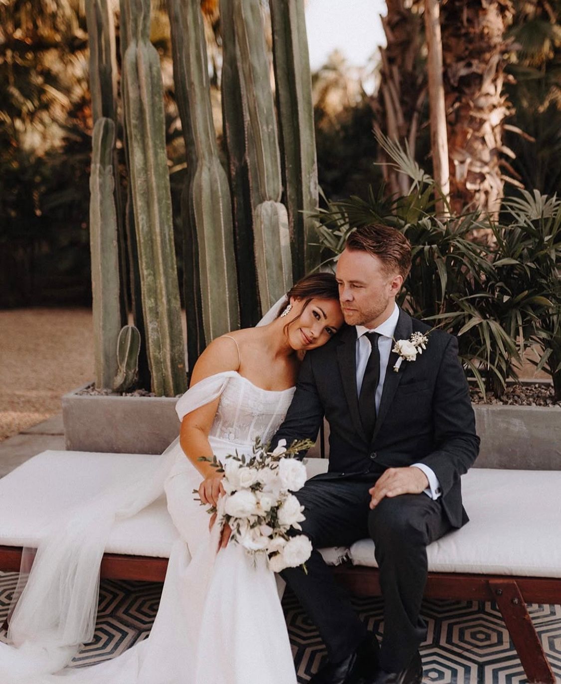 A bride and groom are sitting on a couch in front of a cactus.