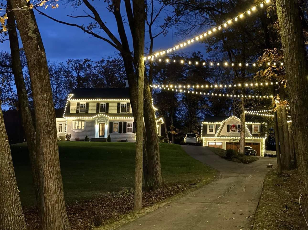 Houses decorated with string lights at dusk. Driveway leads to two homes, trees frame the scene.