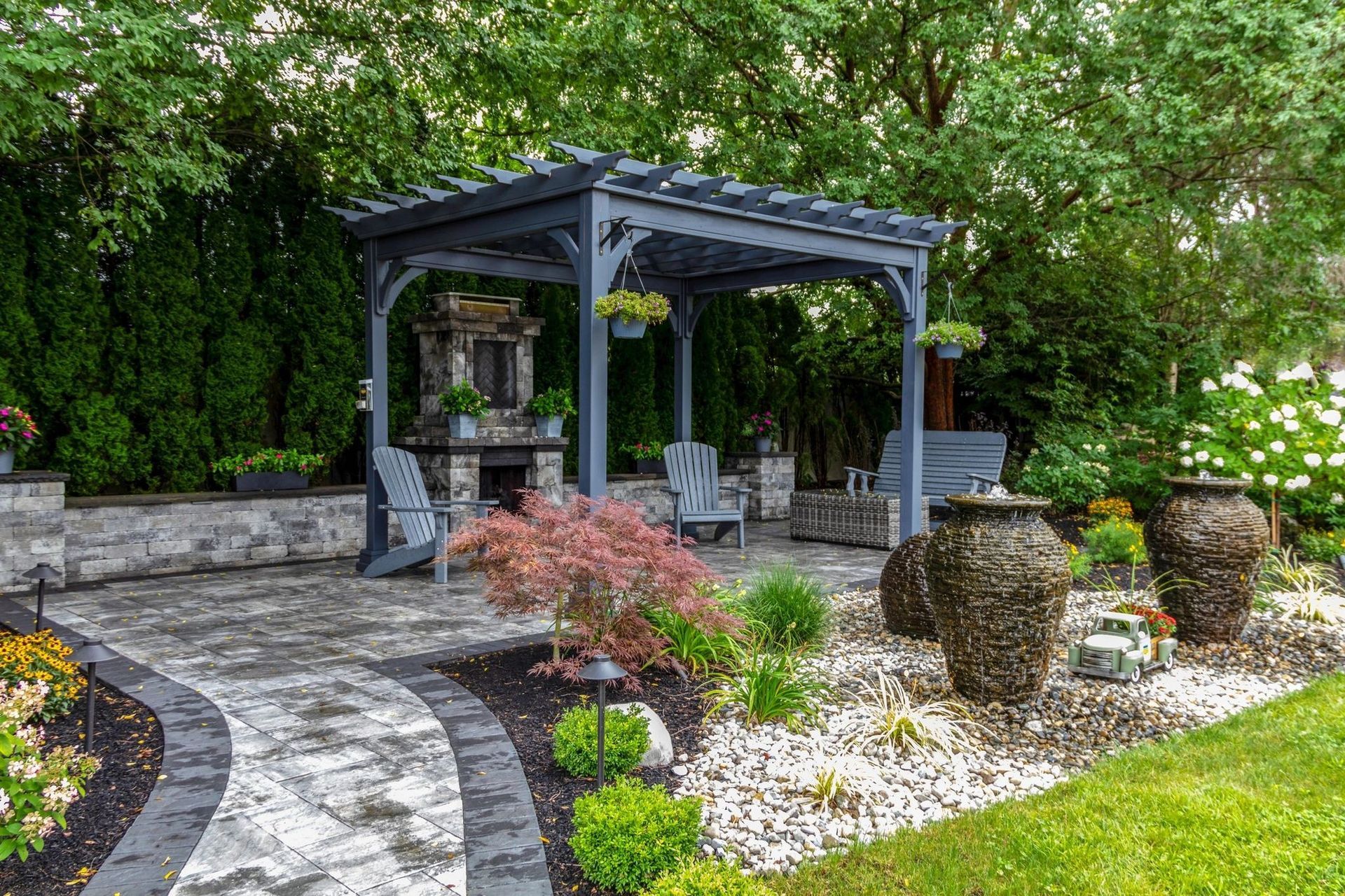 Gray pergola over stone patio with fireplace, seating, and decorative urns in a landscaped backyard.