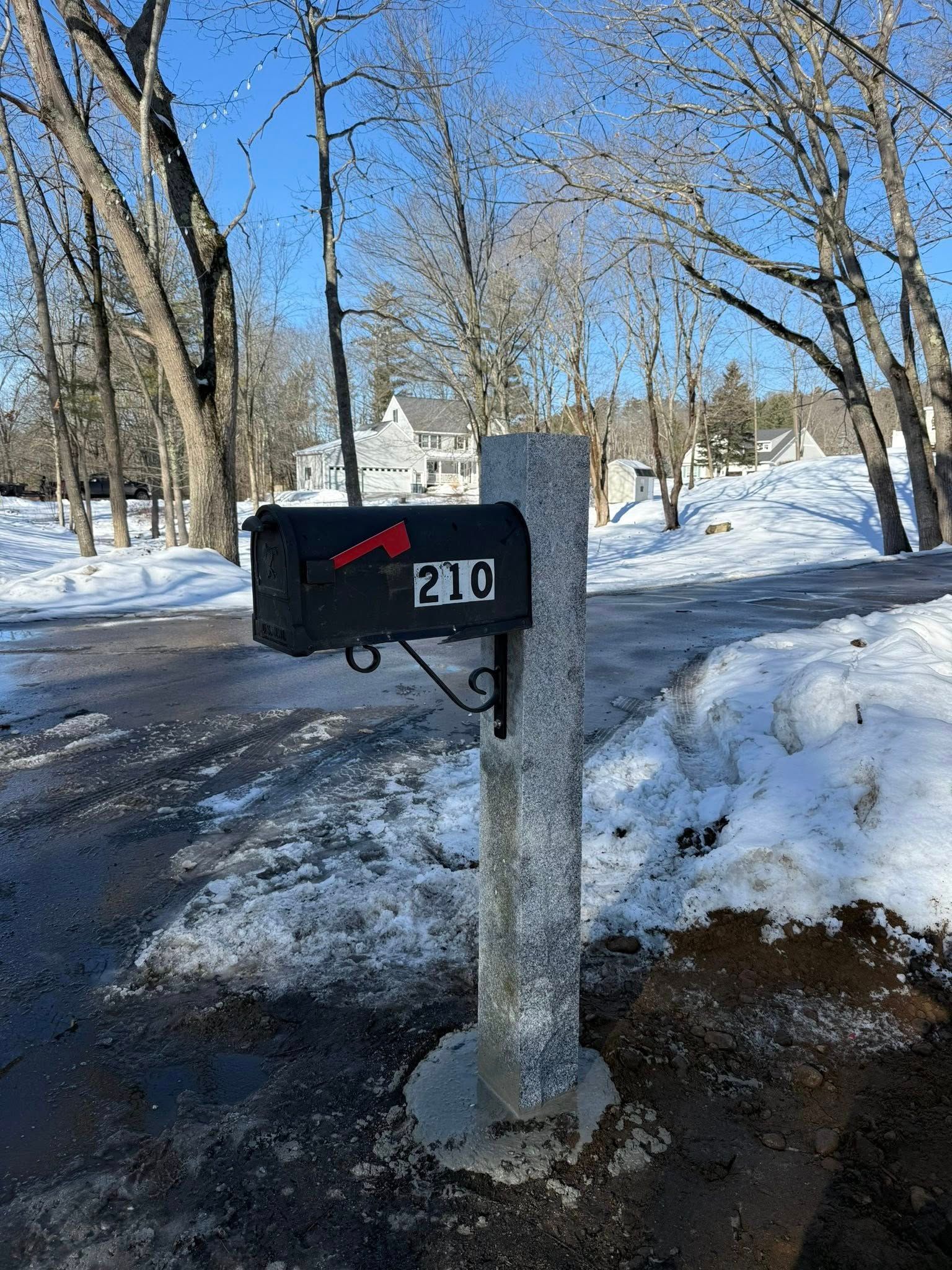 Black mailbox on a concrete post; address 210 in red, snow on ground and trees in background.