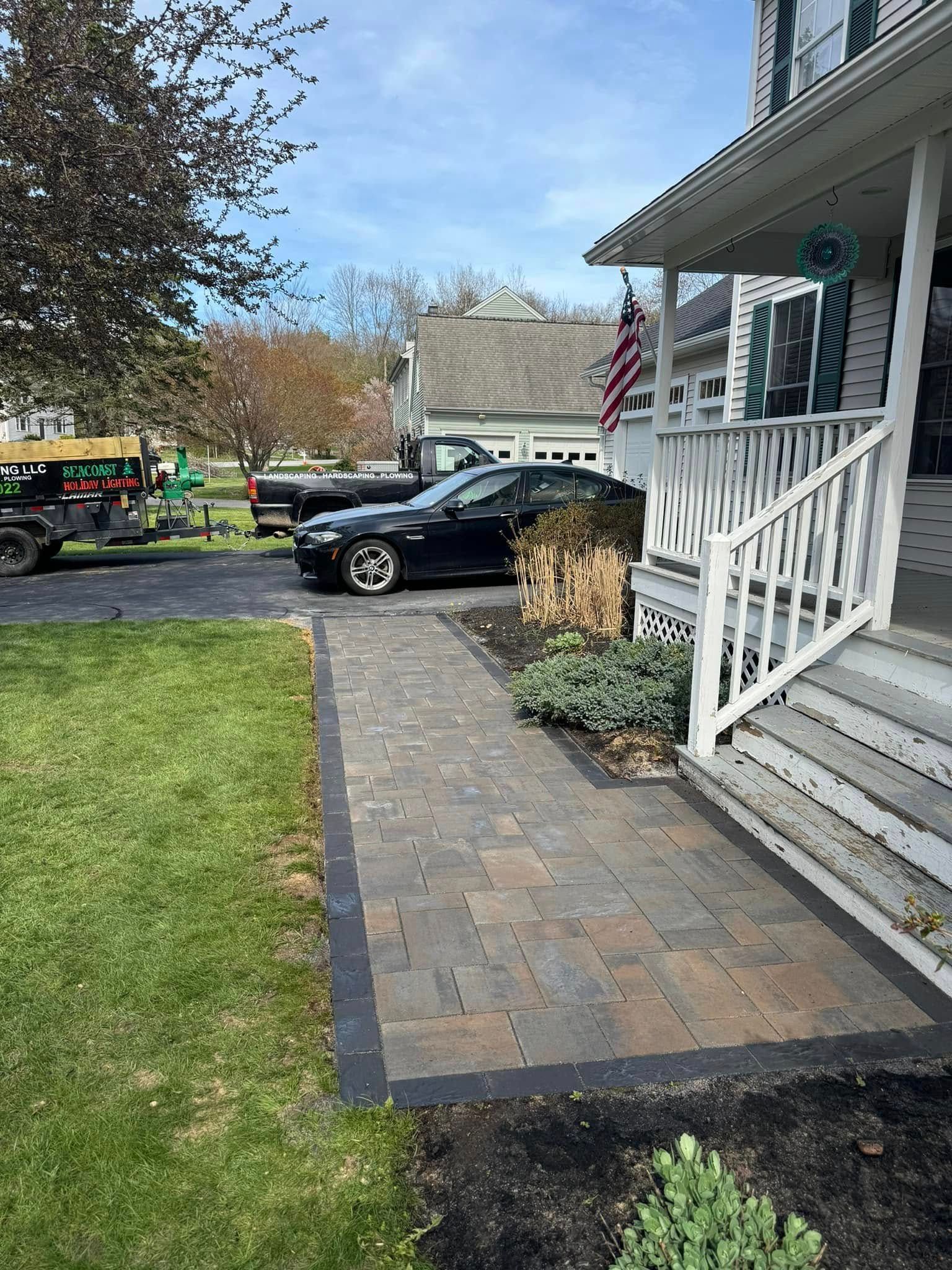 Brick walkway leads to a house porch with American flag; black car parked in driveway.
