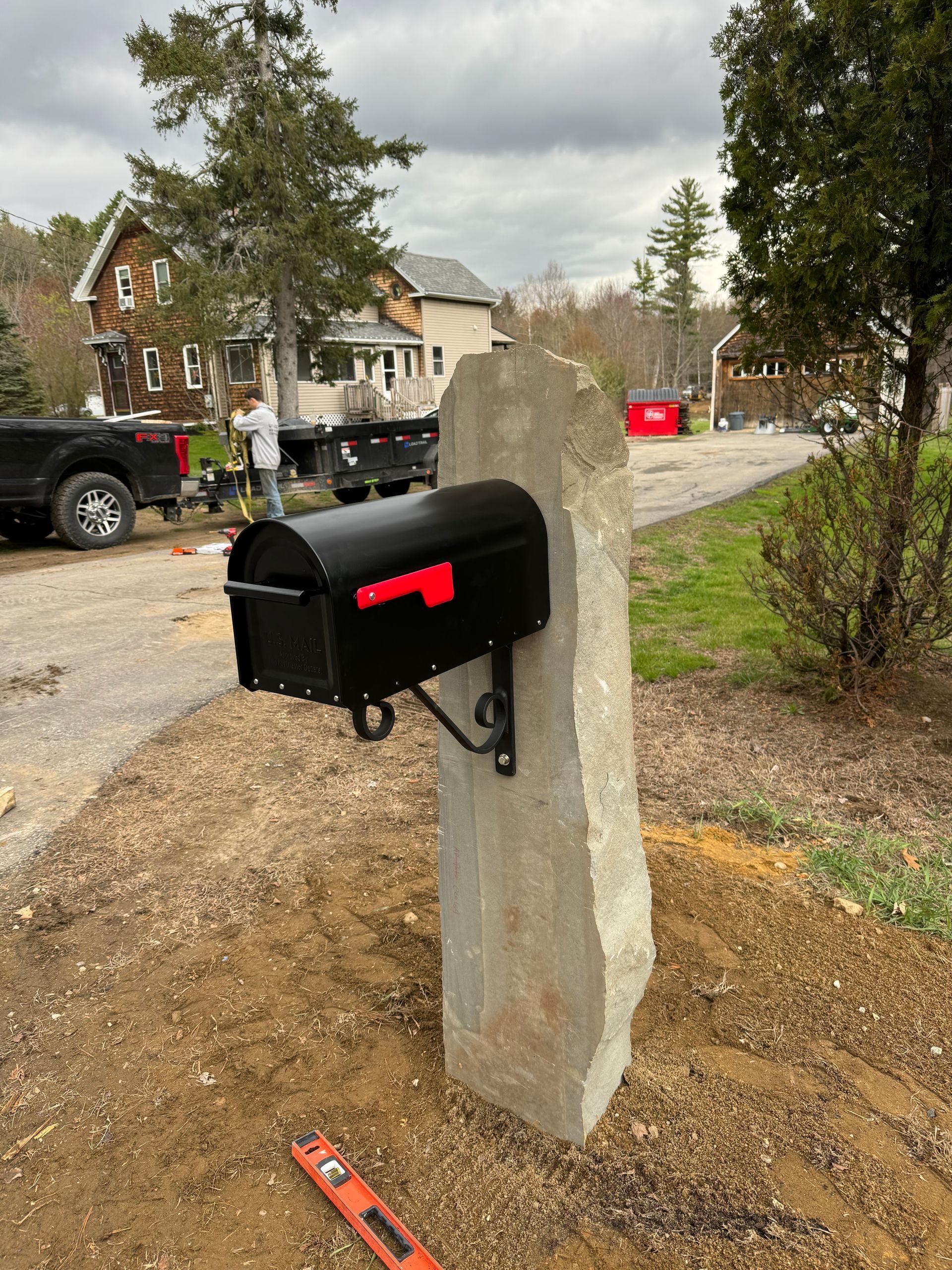 Black mailbox mounted on a concrete post beside a dirt driveway in a suburban neighborhood.