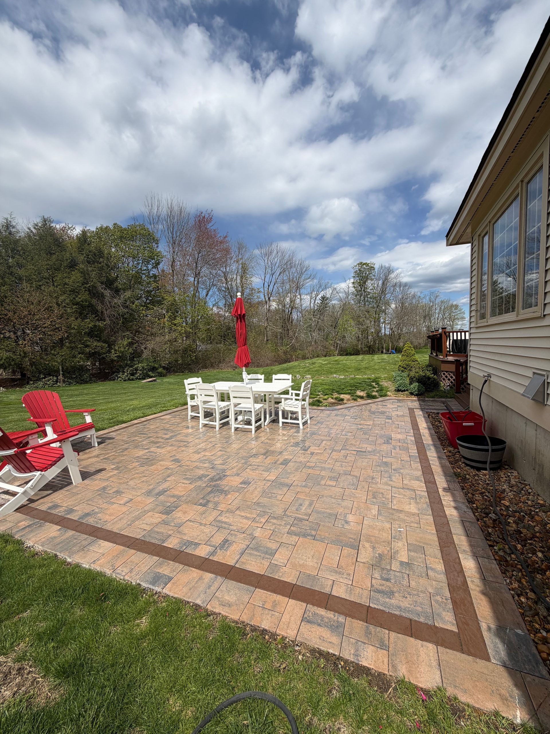 Brick patio with white outdoor dining set, red umbrellas, and lounge chairs beside a grassy yard.