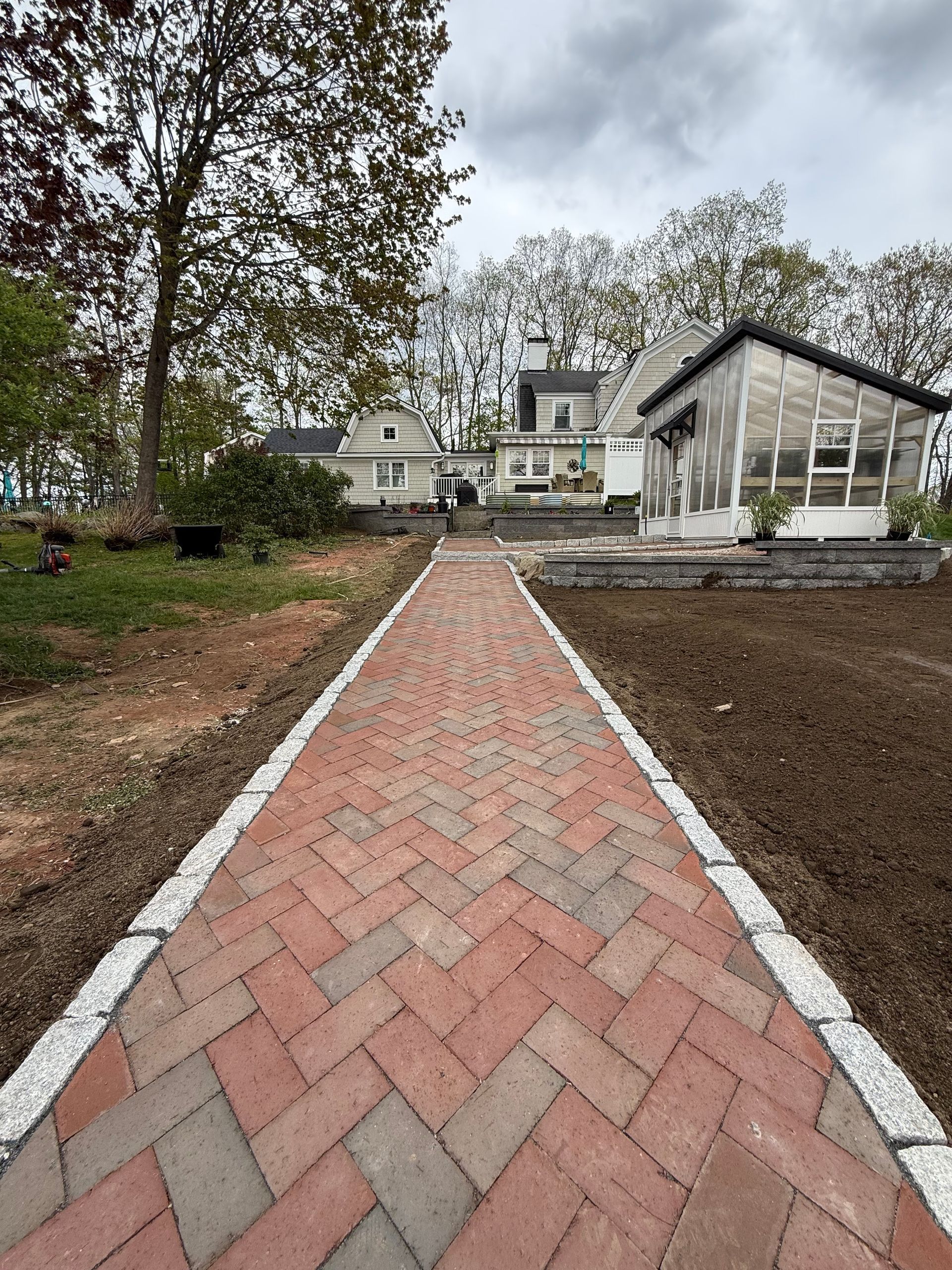 Brick walkway leading to a white house and sunroom, bordered by bare landscaping and trees.