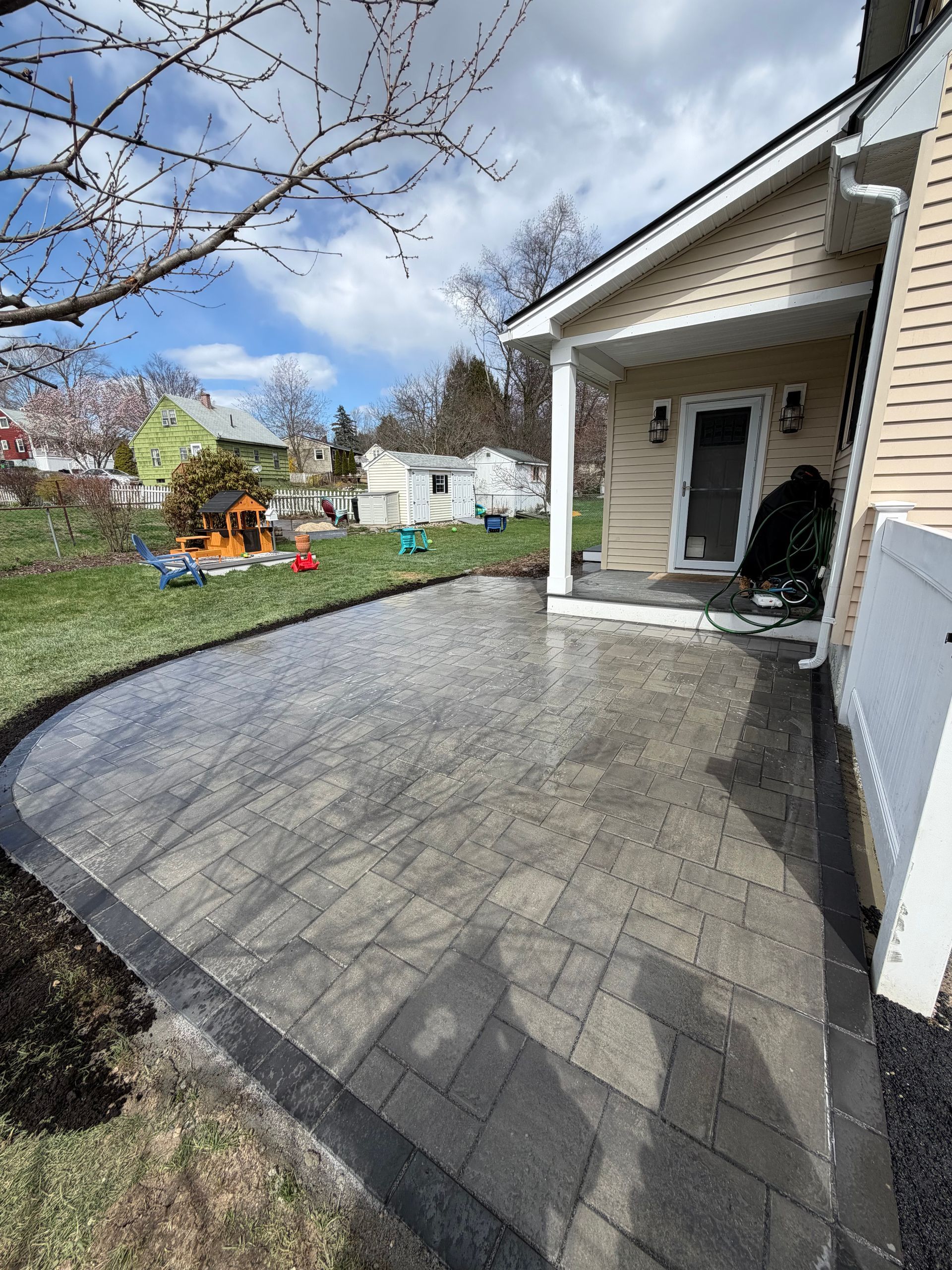 Stone patio beside a white house with a covered porch, overlooking a backyard and blue sky. 